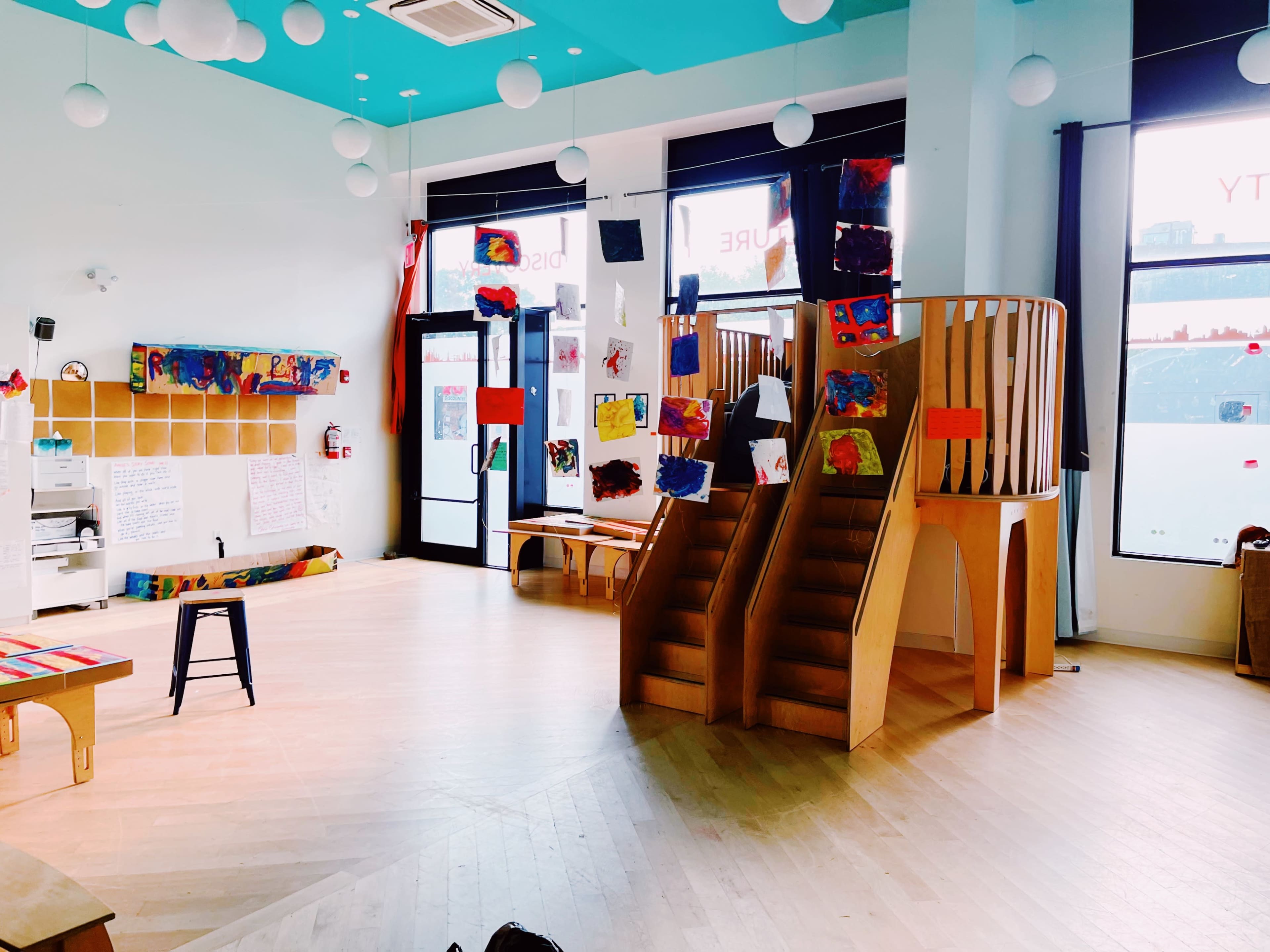 A well-lit classroom featuring a play area with colorful artwork displayed on the walls and a wooden play structure with two slides.