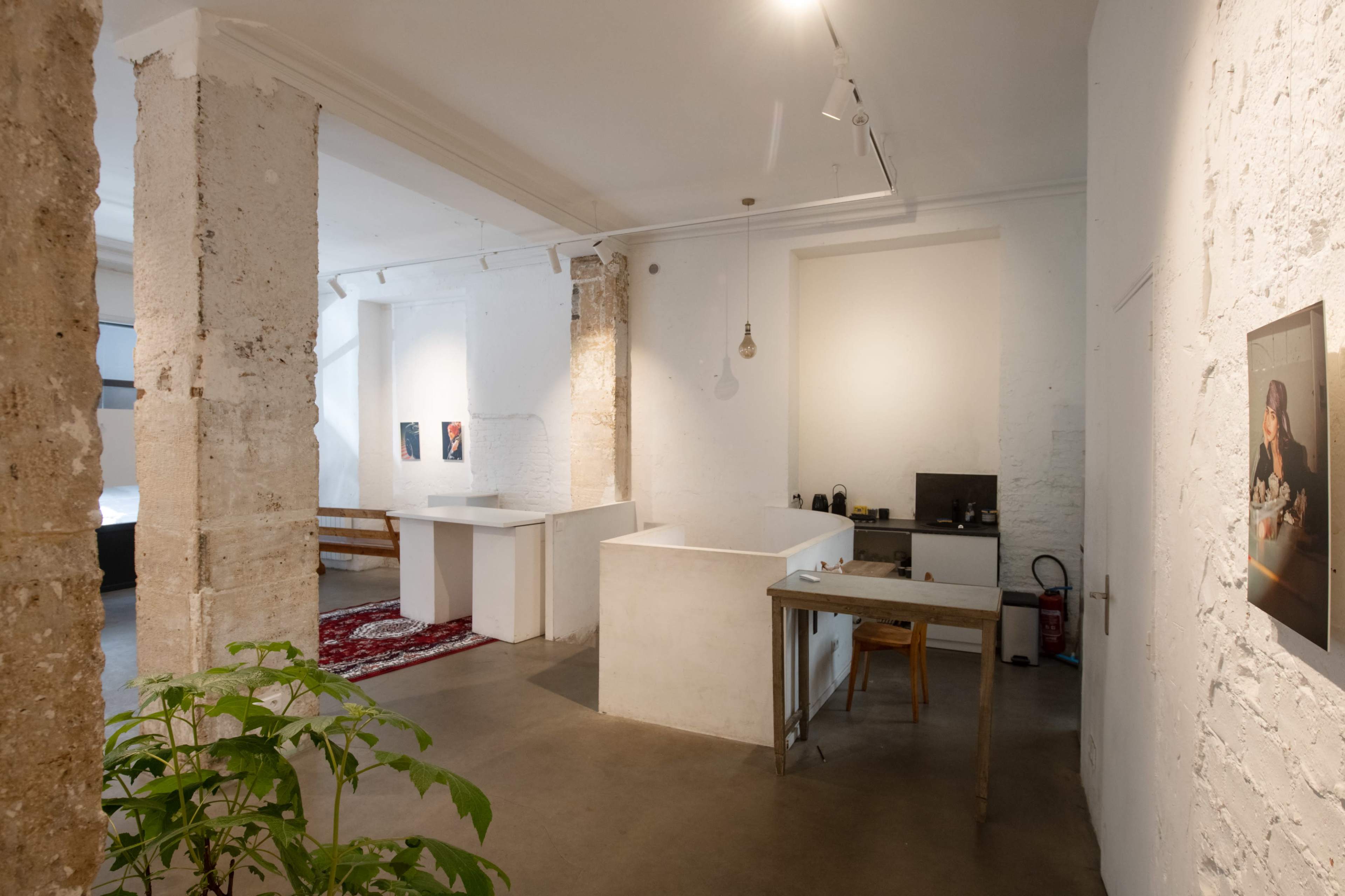 The image shows a minimalist interior space featuring white walls, concrete columns, a wooden table, and a small kitchenette area.