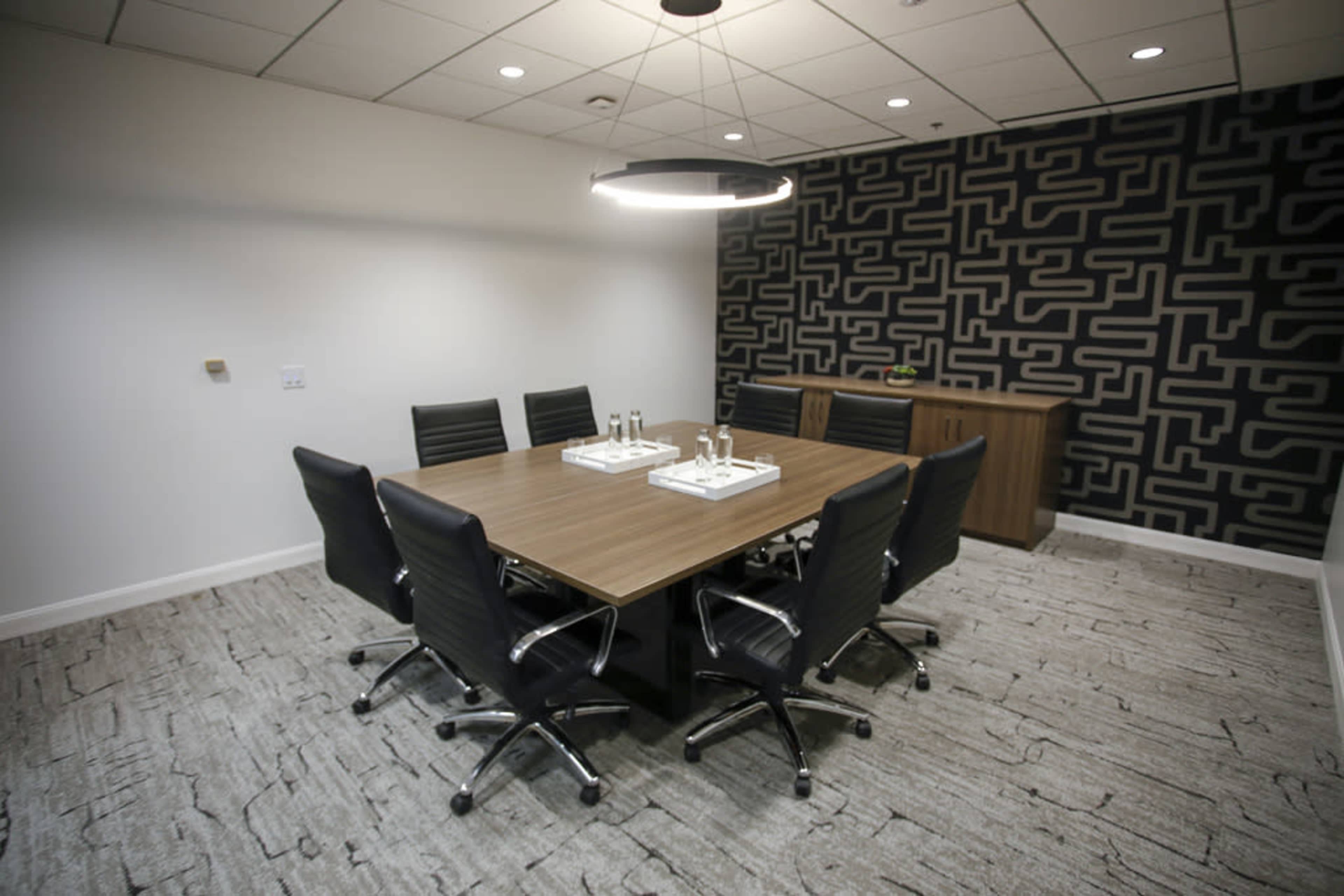 A modern conference room features a large wooden table surrounded by six black swivel chairs, with a patterned accent wall and a side cabinet.