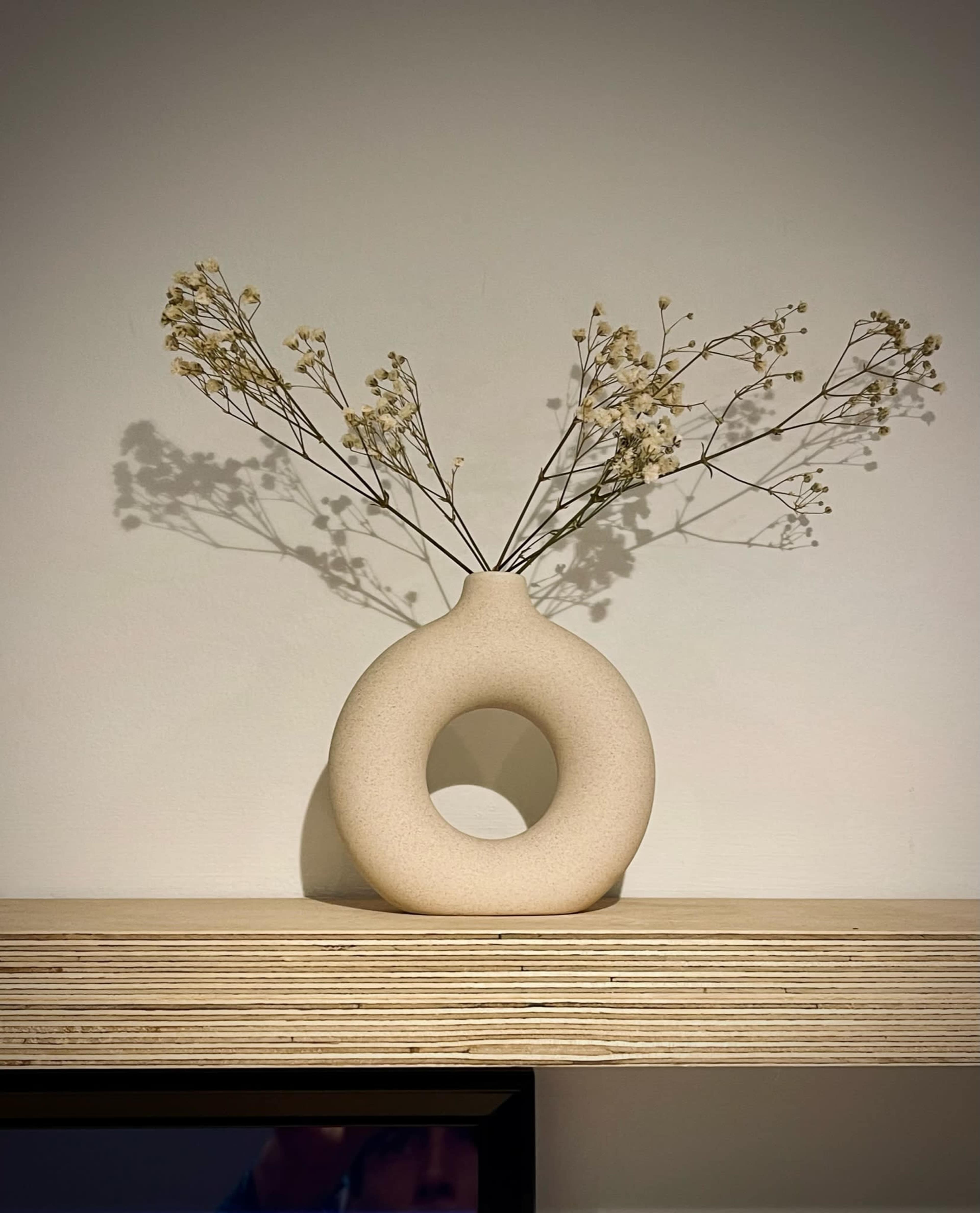 A circular, beige vase with a hole in the center holds dried flowers and is placed on a wooden shelf against a plain wall.