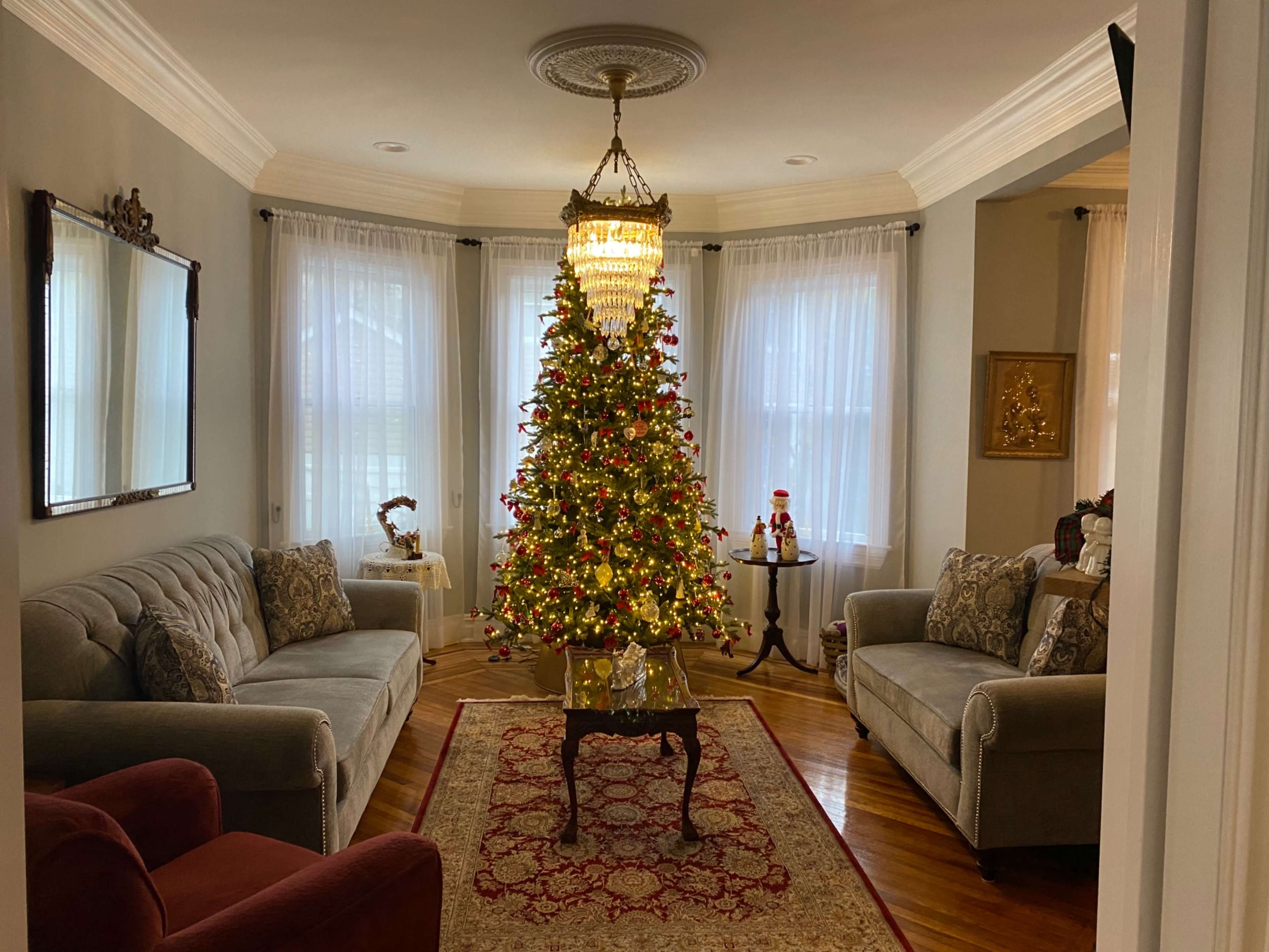 A living room features a decorated Christmas tree in the center, flanked by two sofas and a small table, with large windows allowing natural light to enter.