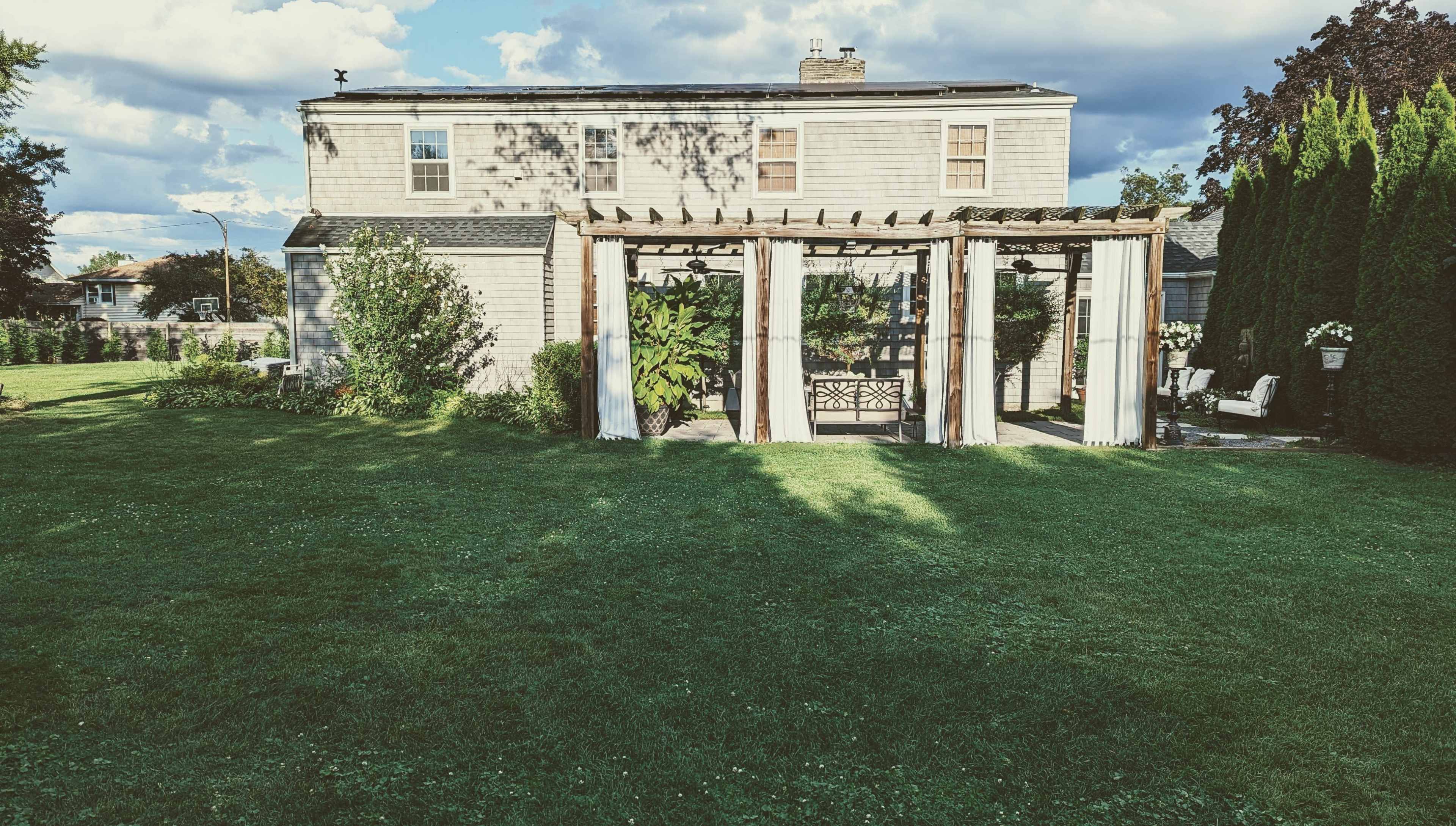 A backyard with a pergola covered in light drapes, grass, and a house in the background under a partly cloudy sky.