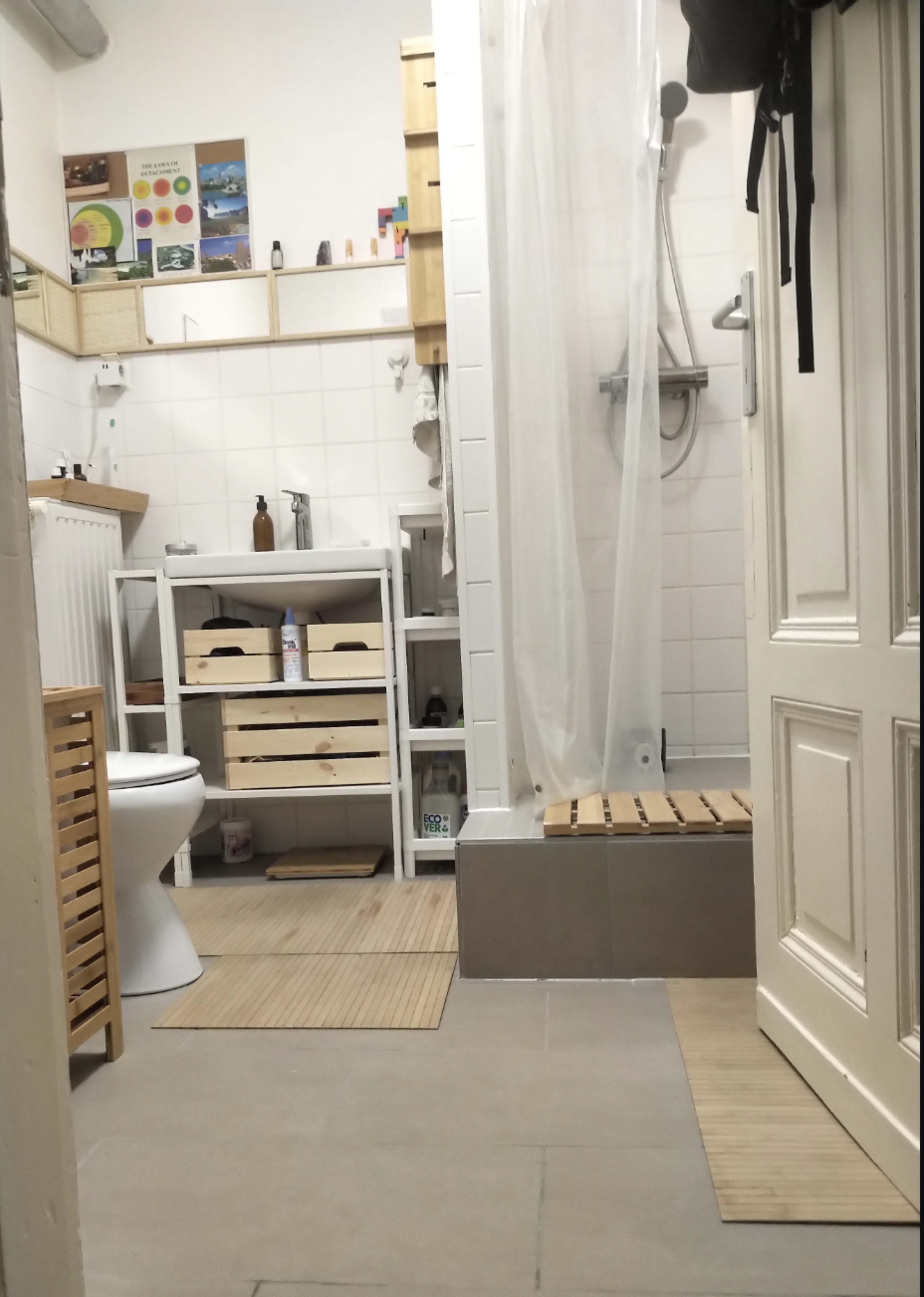 The image shows a compact bathroom with a shower, toilet, and wooden shelving units, featuring light-colored tiles and a curtain separating the shower area.