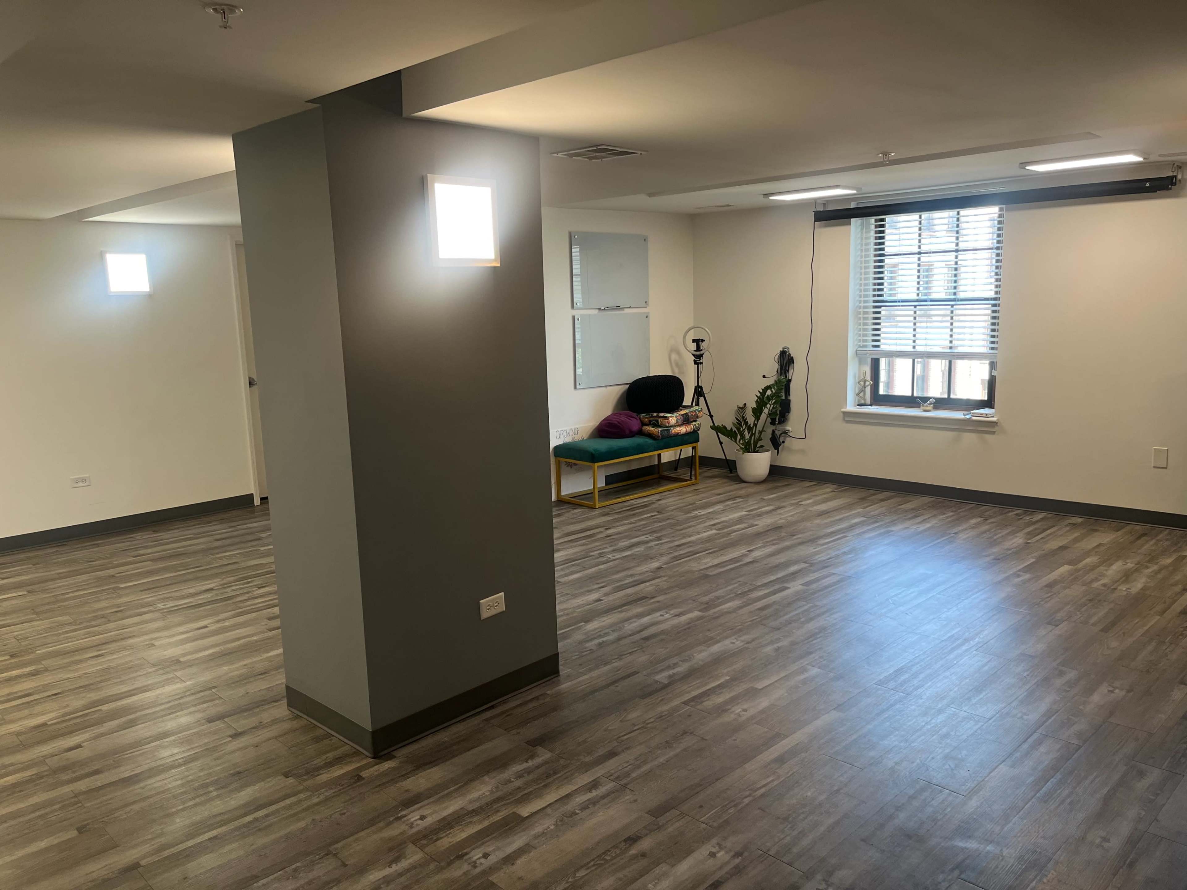An empty interior room with wood-like flooring, a few windows, and minimal furniture including a bench and a plant.