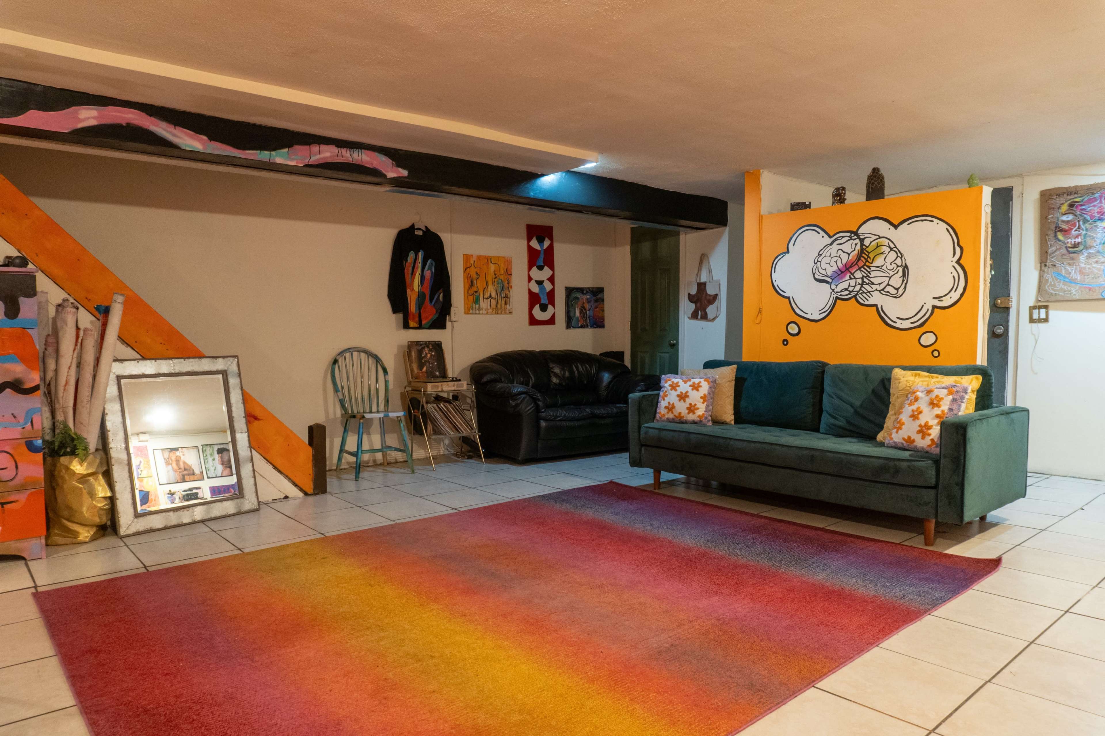 The scene shows a colorful basement lounge area with a green couch, a black leather sofa, a large area rug in gradient colors, and various artworks on the walls.