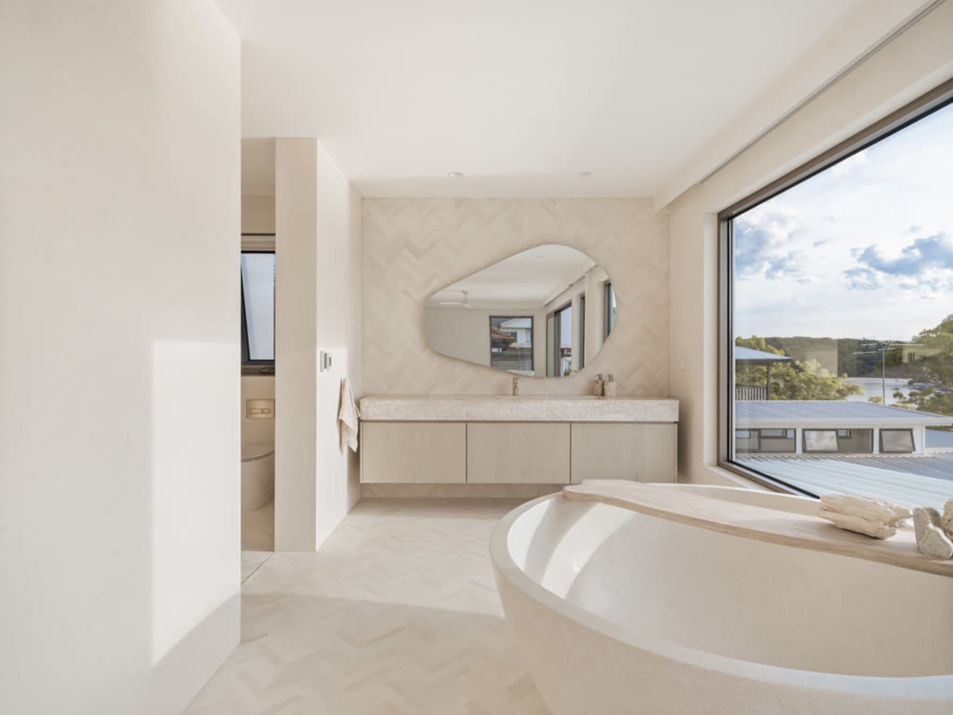 The image shows a modern bathroom featuring a freestanding bathtub, a large mirror above a sleek vanity, and a large window with a view of nature.