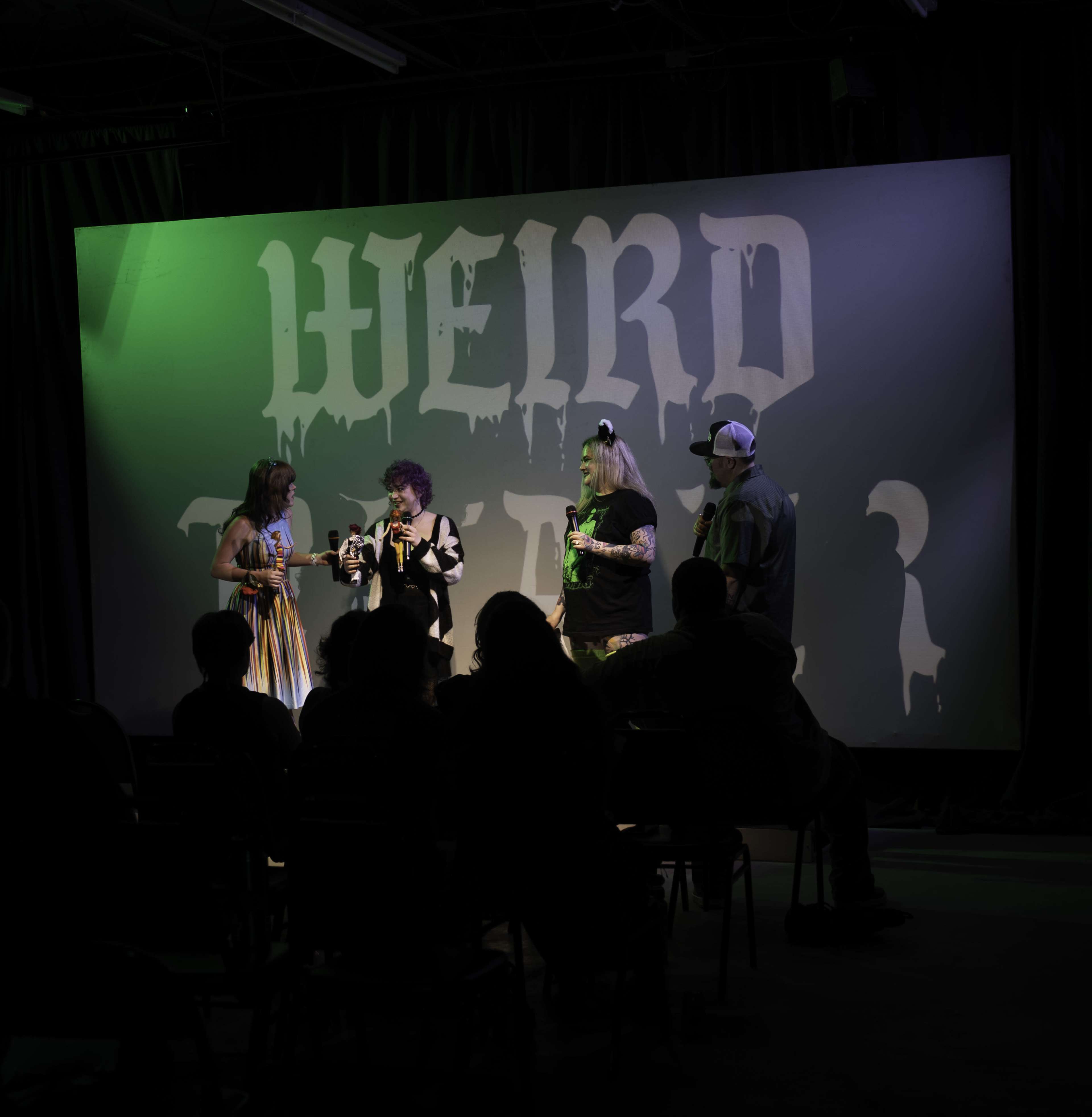 A group of four performers stands on stage in front of a backdrop that reads "WEIRD" while an audience watches from their seats.