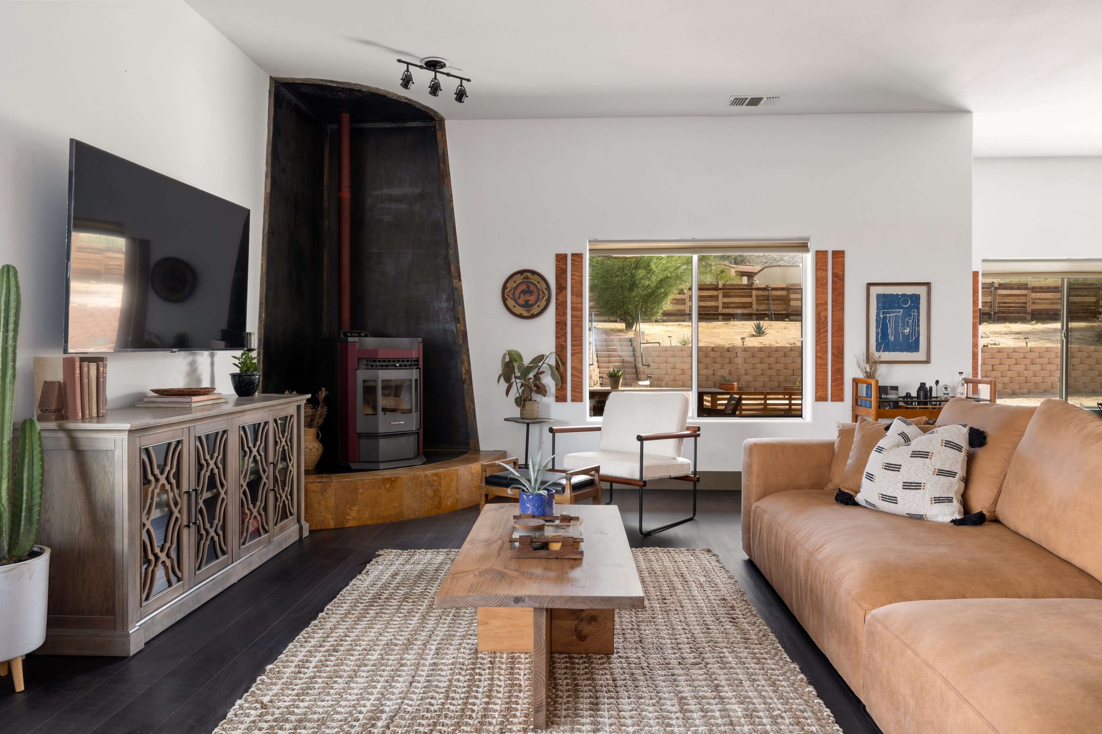 The living room features a large sectional couch, a wooden coffee table, and a modern television mounted on the wall, with a cozy wood stove in the corner and windows showcasing an outdoor view.
