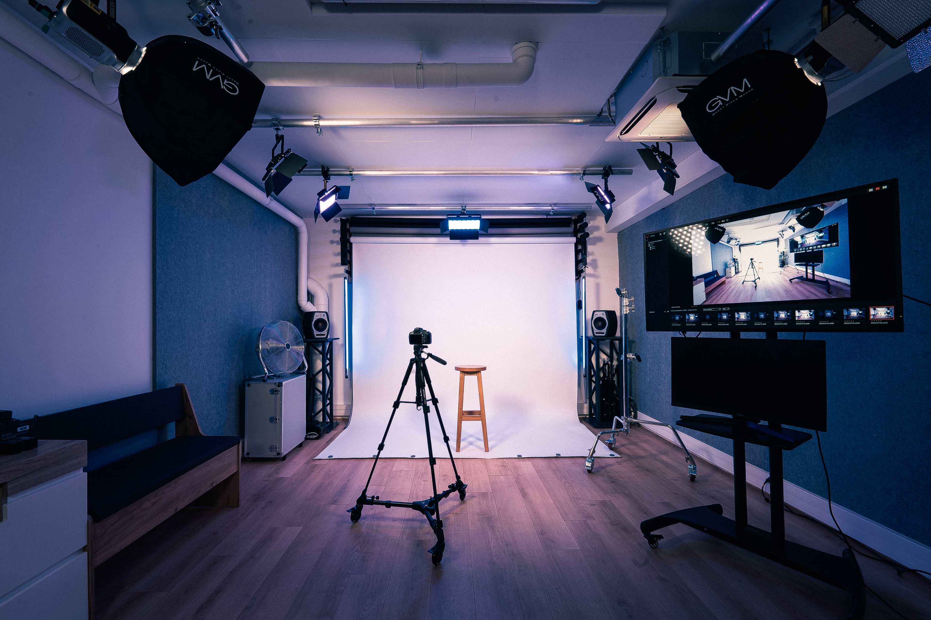 A photography studio is set up with a camera on a tripod facing a wooden stool and a white backdrop, illuminated by multiple studio lights.