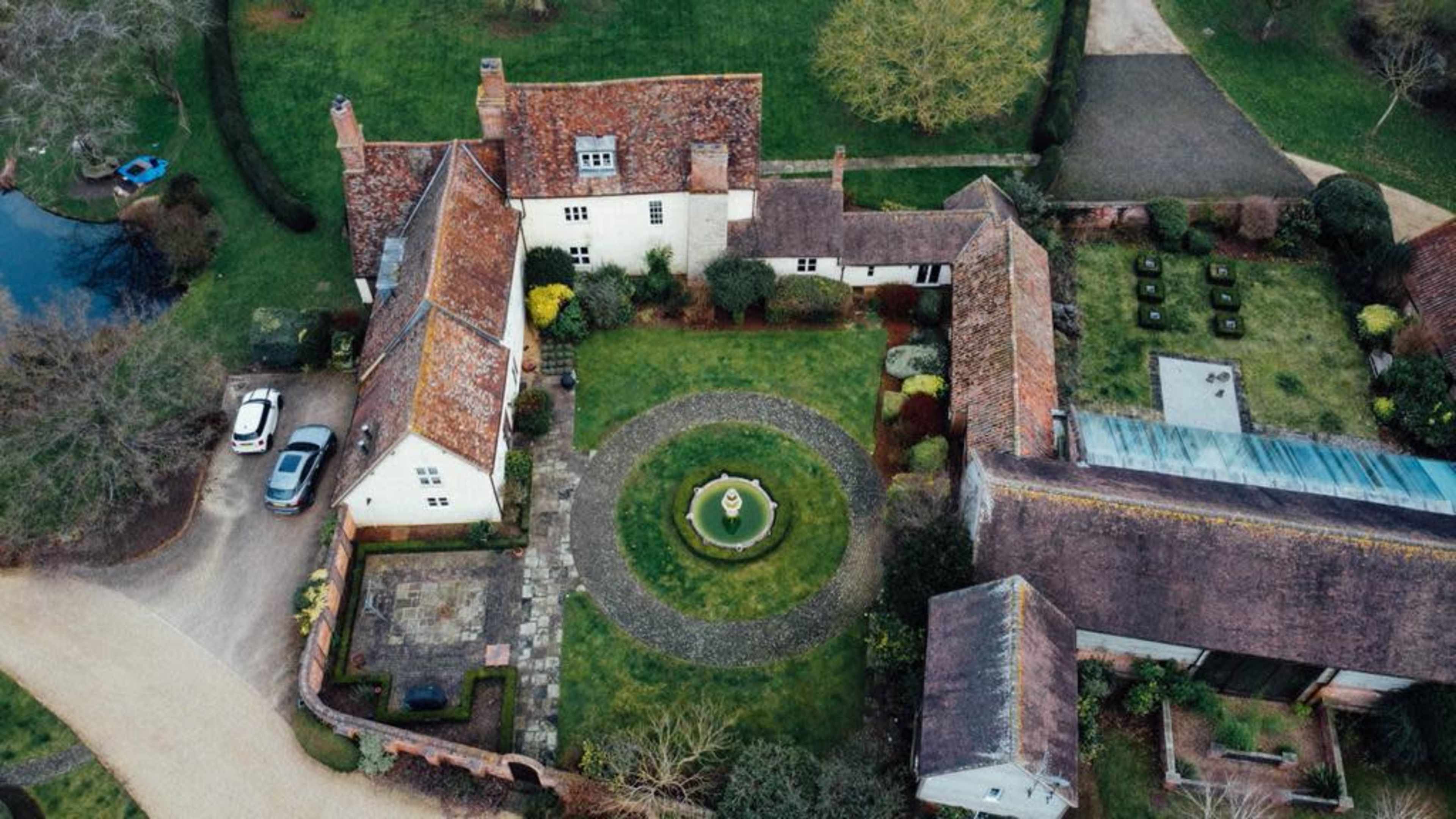 An aerial view of a landscaped property featuring a circular fountain in the center of a garden surrounded by multiple buildings and a car parked nearby.