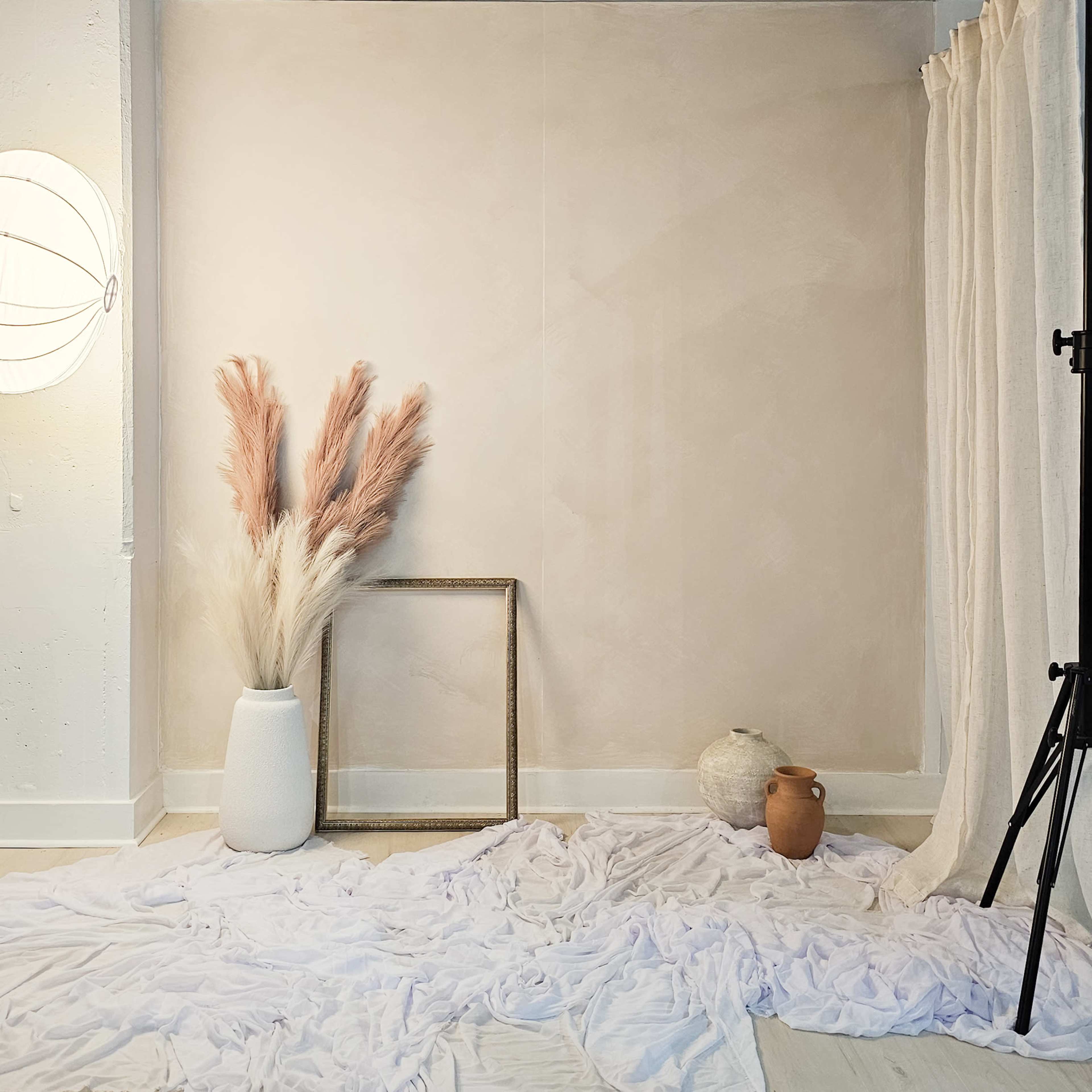 A minimalist indoor scene features a textured wall, a vase with dried pampas grass, an empty frame, and a few decorative objects arranged on a soft fabric-covered floor.