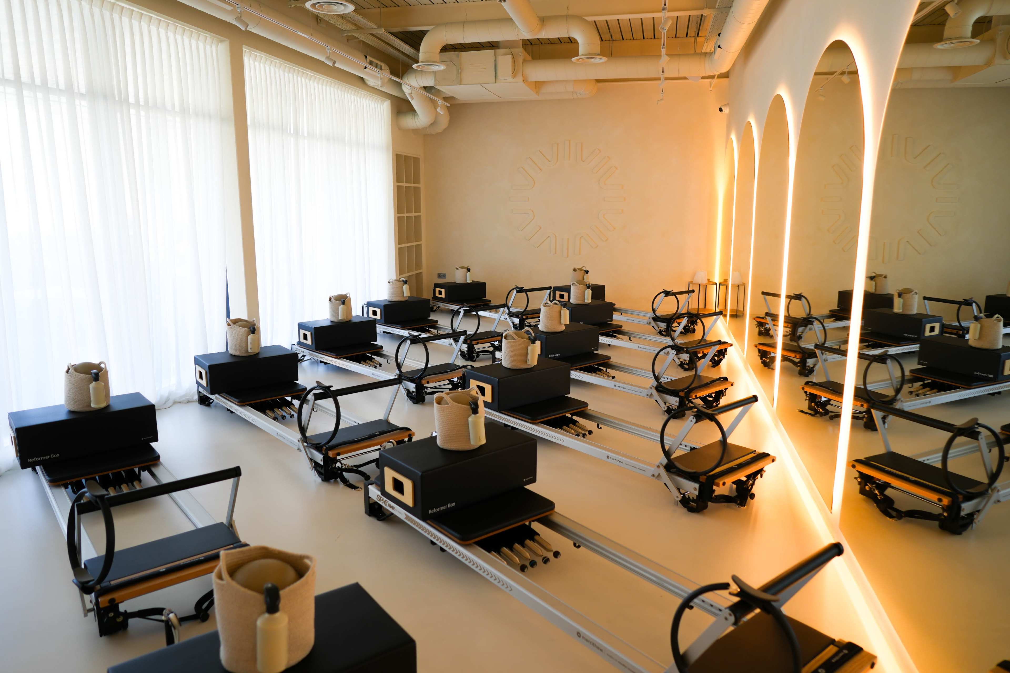 The image shows a spacious fitness studio with rows of pilates reformers arranged neatly in front of large windows and softly lit walls.