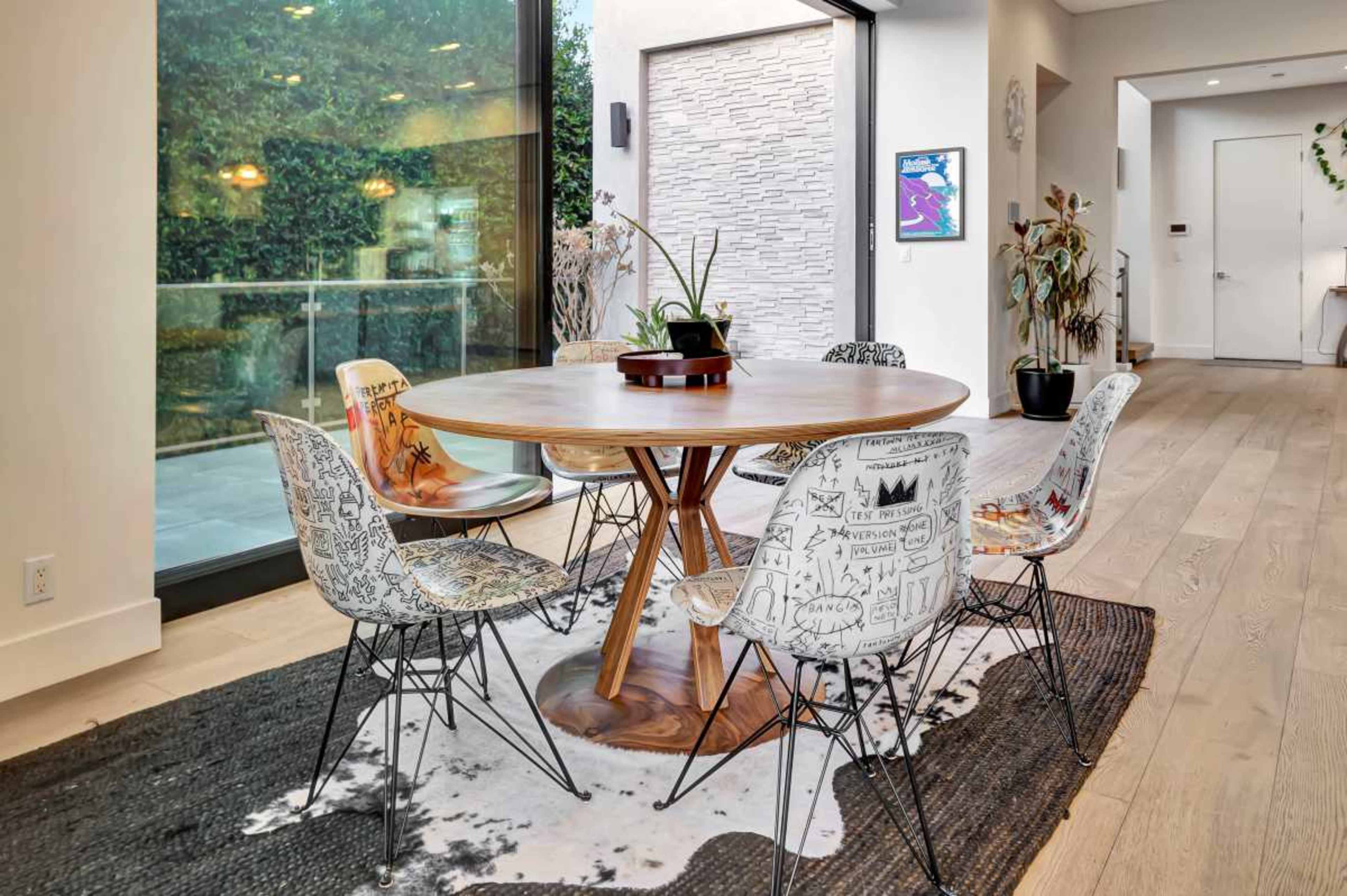The image shows a modern dining area featuring a round wooden table surrounded by artistic chairs, with a large glass door leading to an outdoor space.