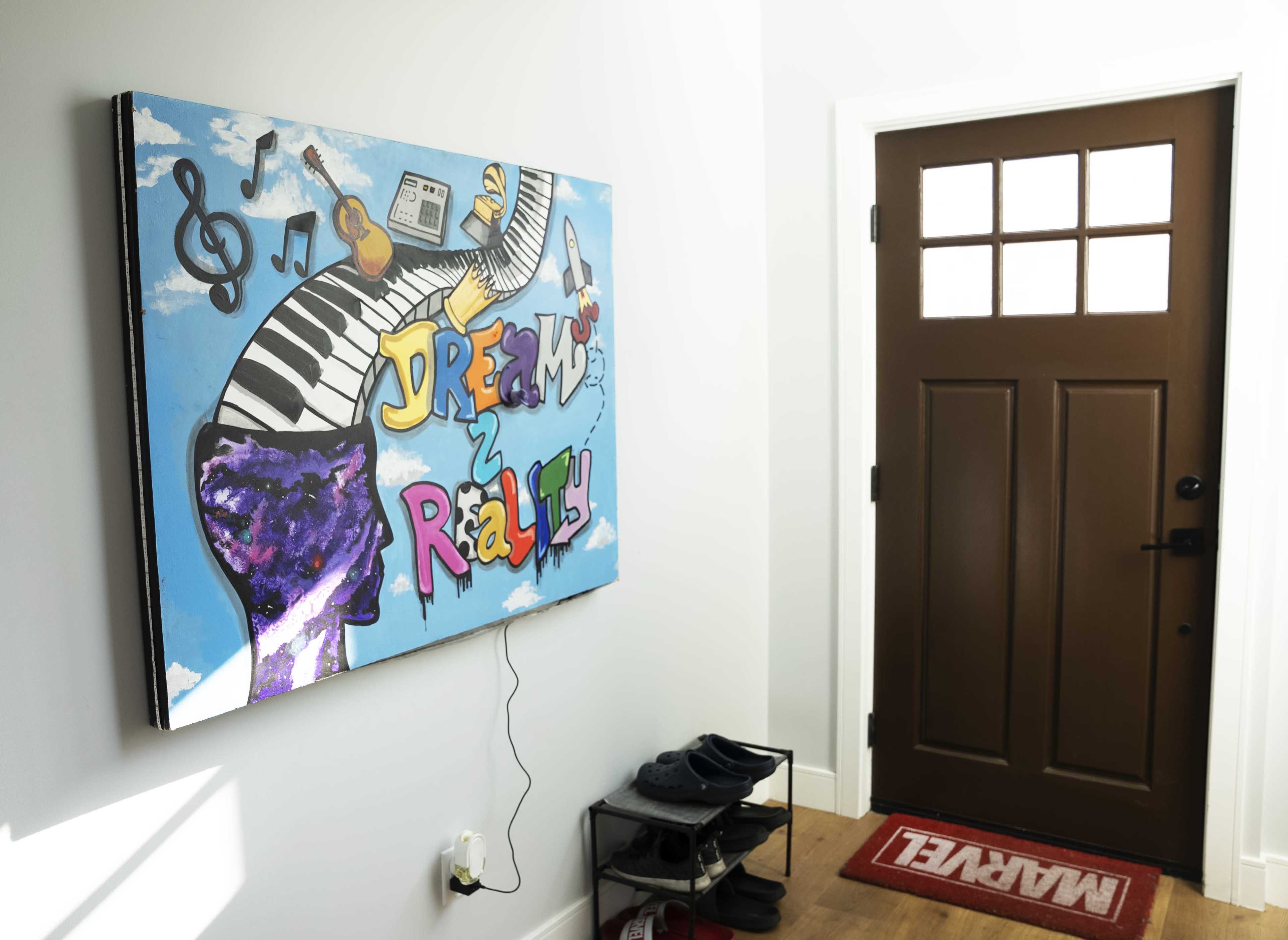 A colorful painting titled "Dreams to Reality" featuring musical elements, displayed on a wall next to a dark wooden door and a welcome mat.