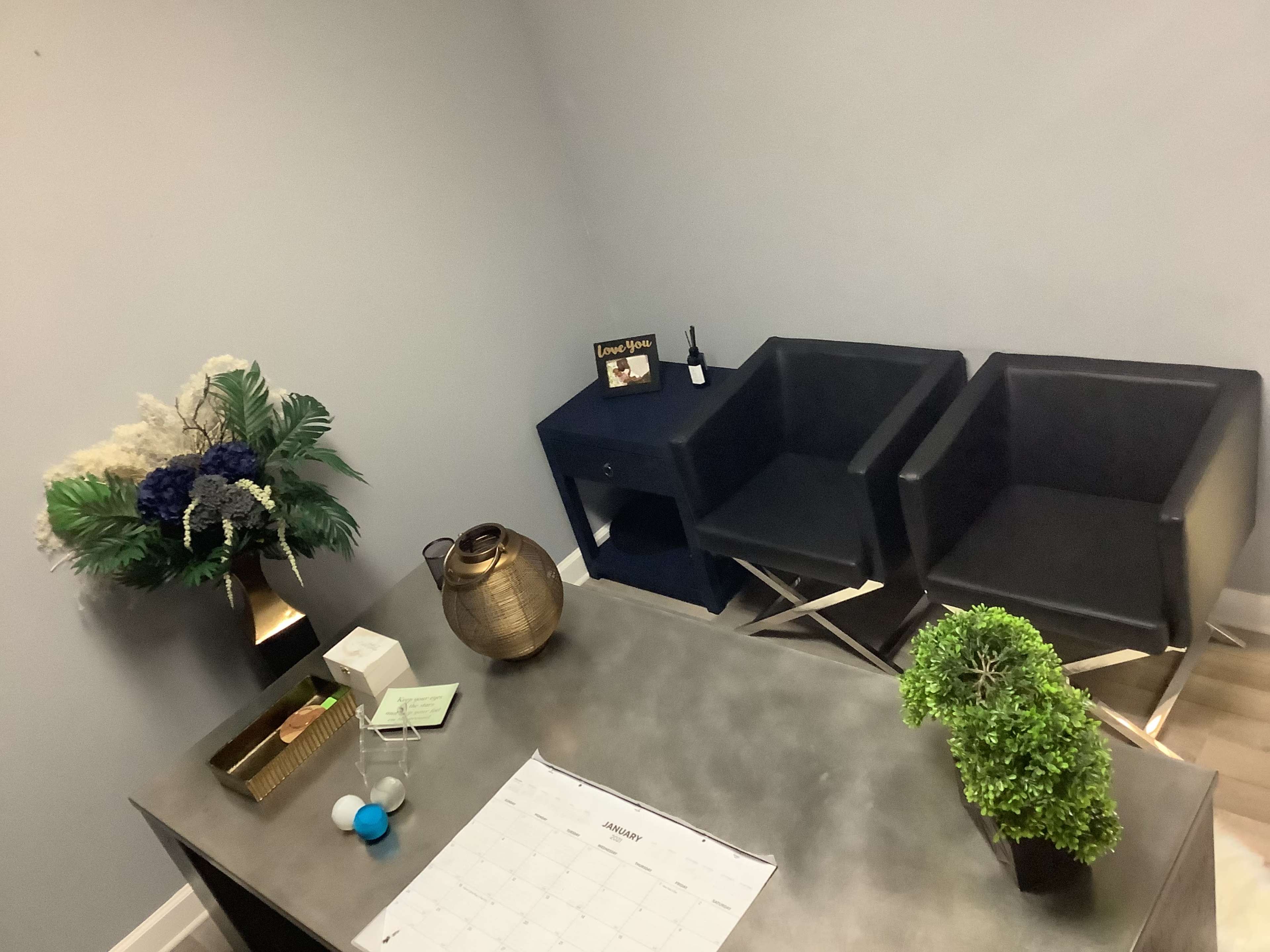 The image shows a small office space with two black chairs, a desk with a calendar, and decorative items including a vase of flowers and a small plant.