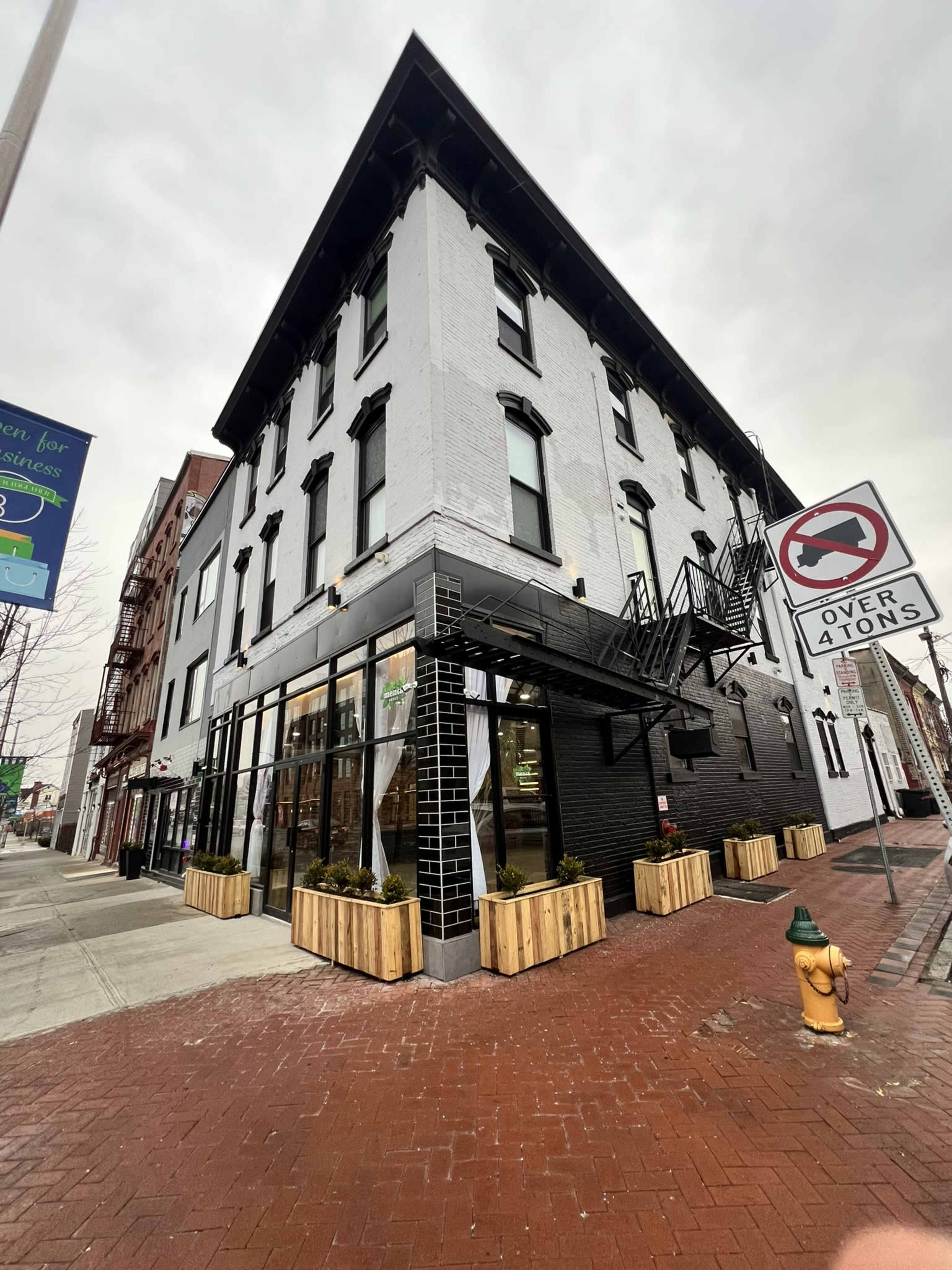 A three-story brick building with large windows is situated on a corner, featuring a black facade, a fire escape, and planters along the sidewalk.