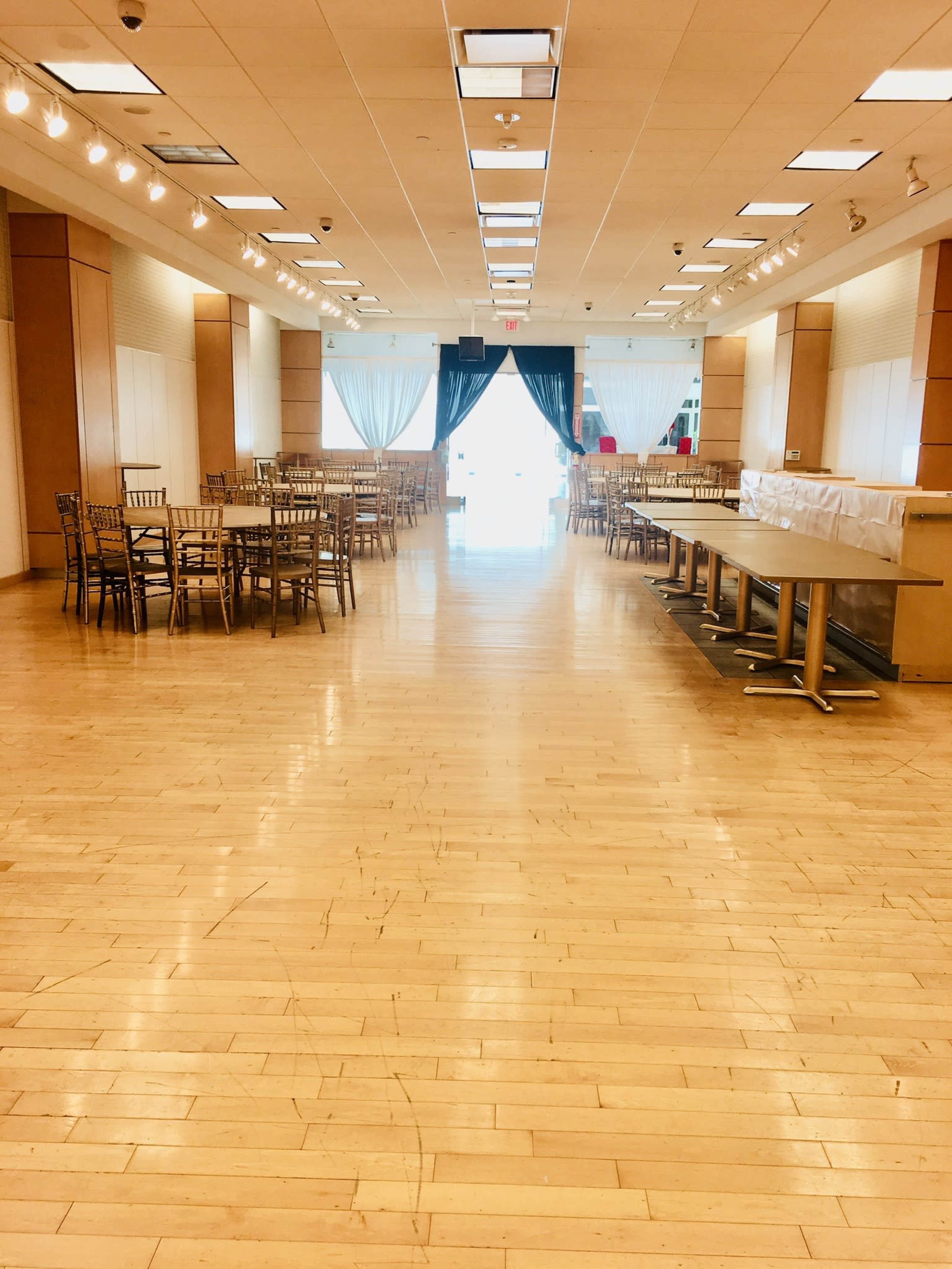 A spacious, empty event hall with wooden floors, arranged with tables and chairs on either side, leading to a bright entrance at the far end.