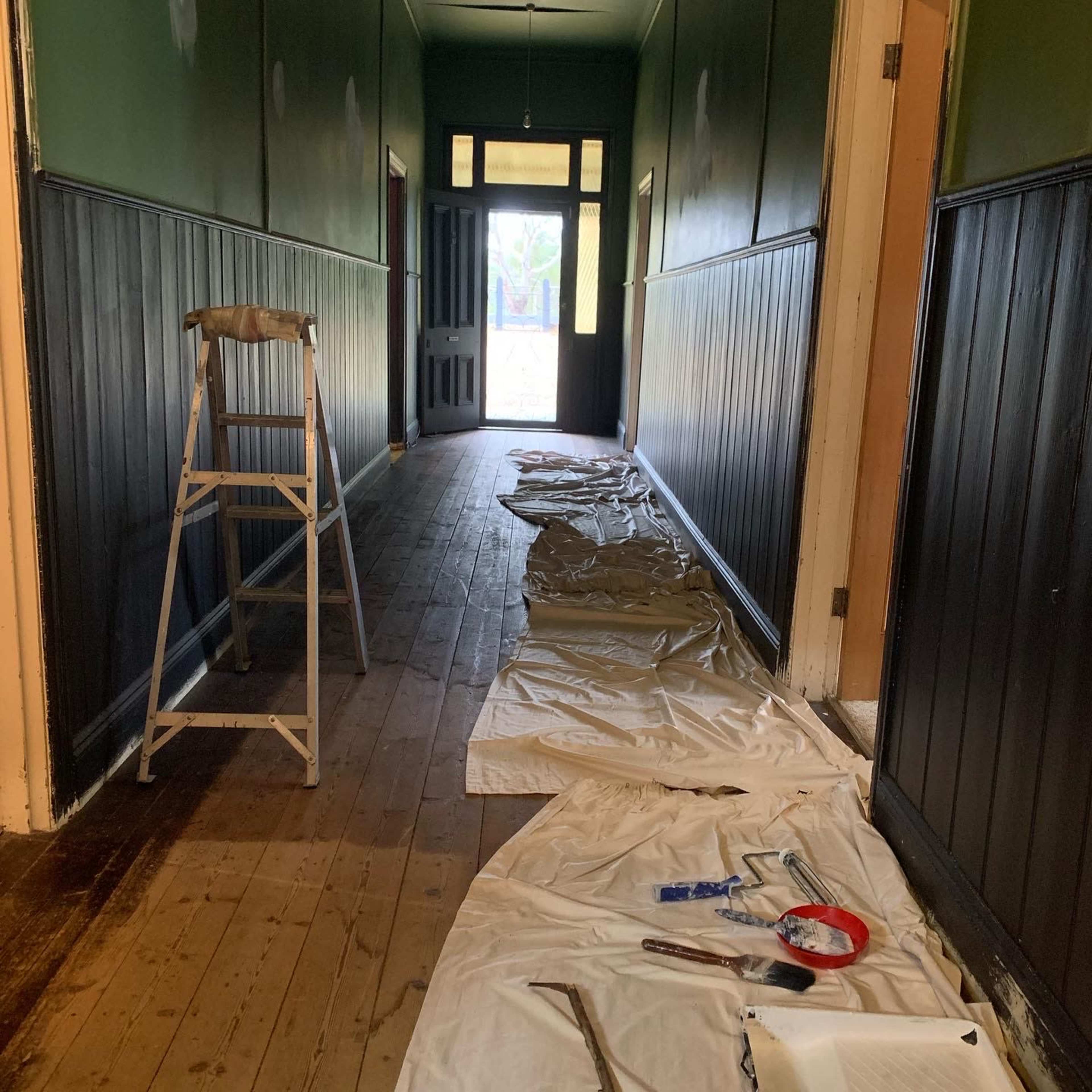 A long hallway with dark wooden floors is covered by drop cloths, and a step ladder is placed to the side, while painting supplies are scattered along the floor.