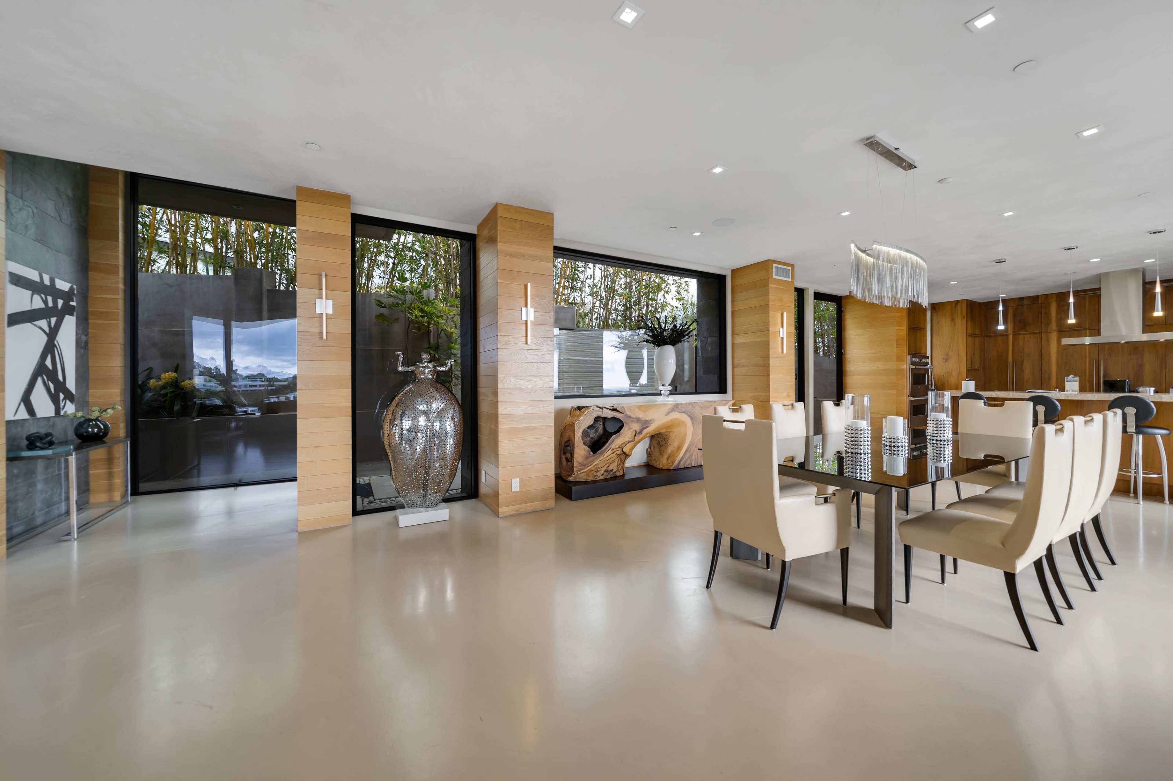 The image shows a modern dining area featuring a large table surrounded by beige chairs, a decorative vase, and large windows that provide a view of an outdoor space.