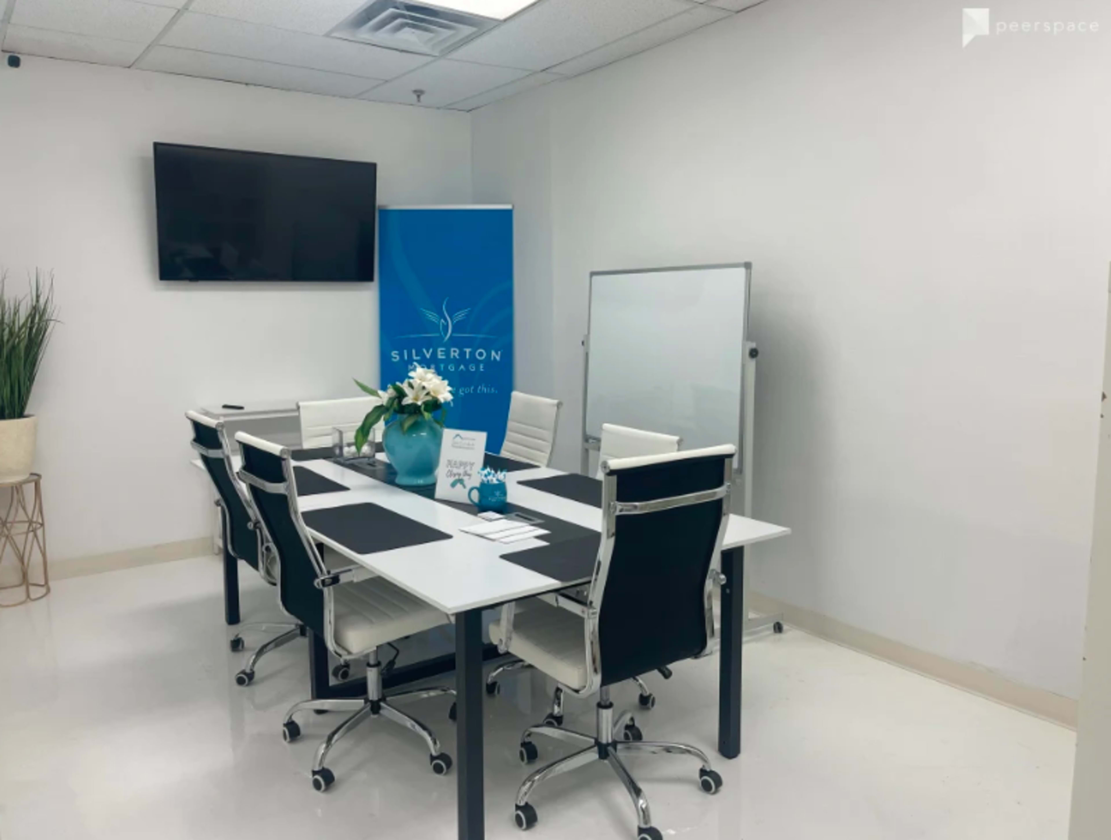 A modern conference room features a large table surrounded by white chairs, a television on the wall, and a plant in the corner.