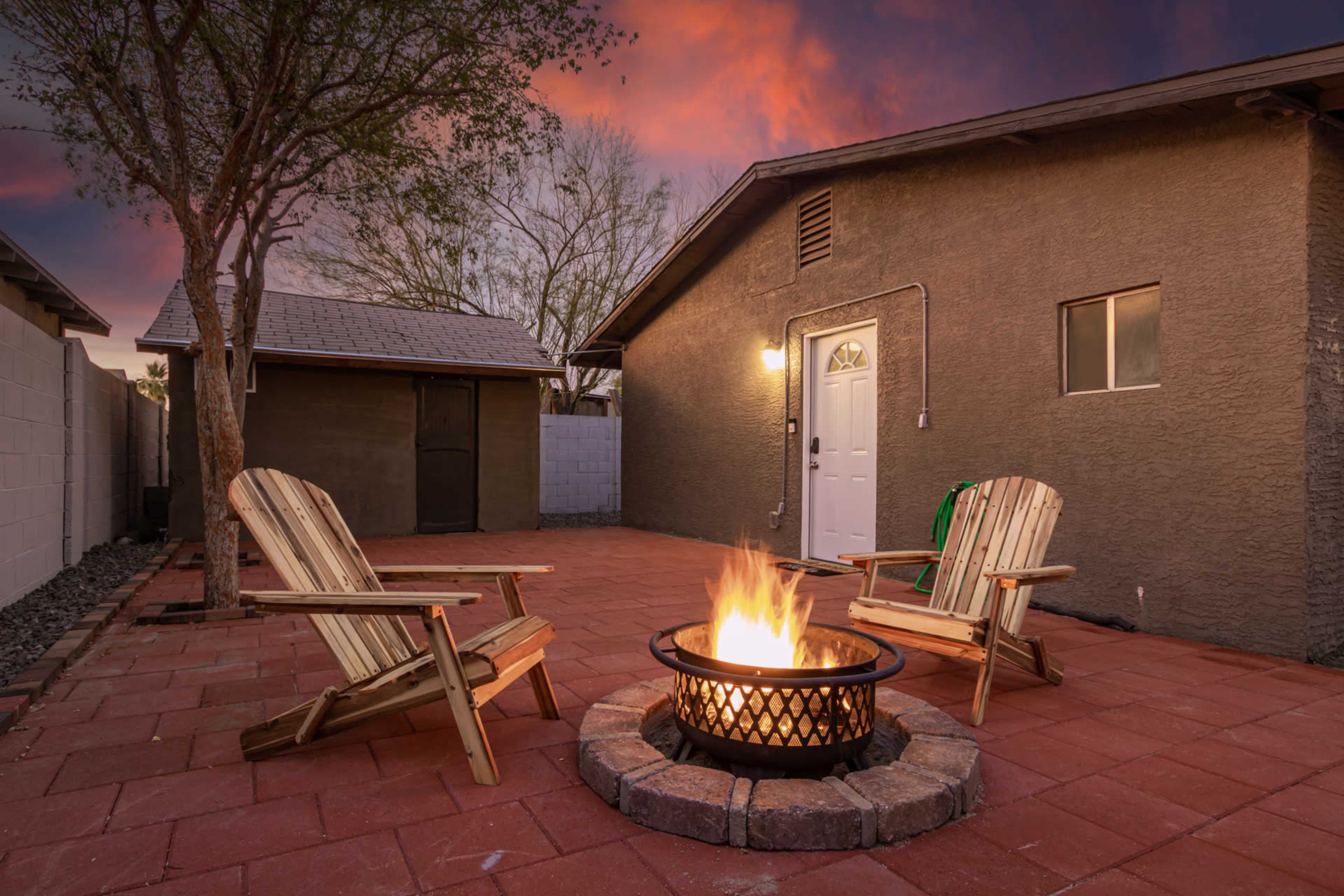 A circular fire pit with flames sits between two wooden lounge chairs on a brick patio, adjacent to a square building at dusk.