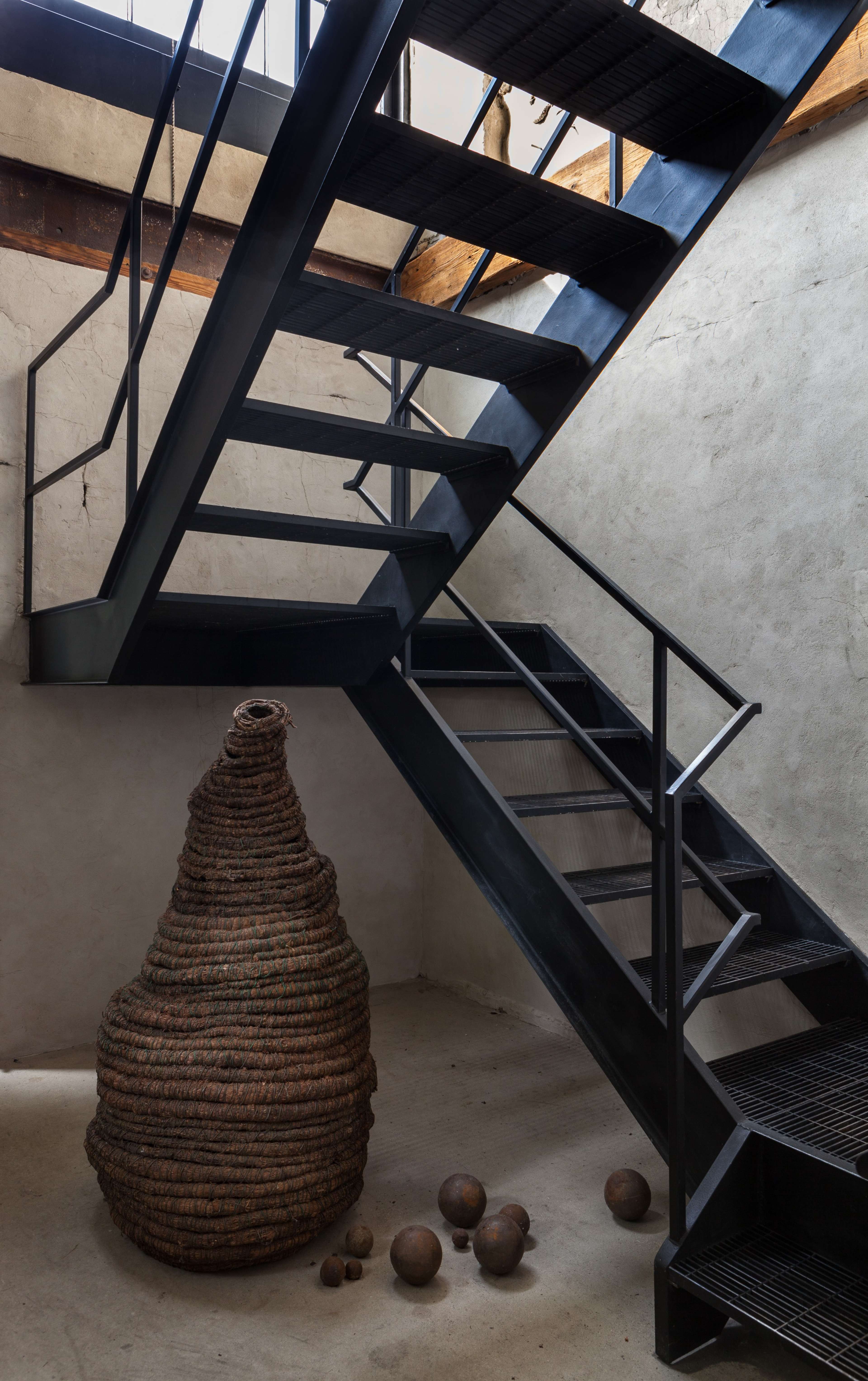 A black metal staircase ascends alongside a textured wall, with a large woven sculpture and several smaller round objects positioned on the floor below.