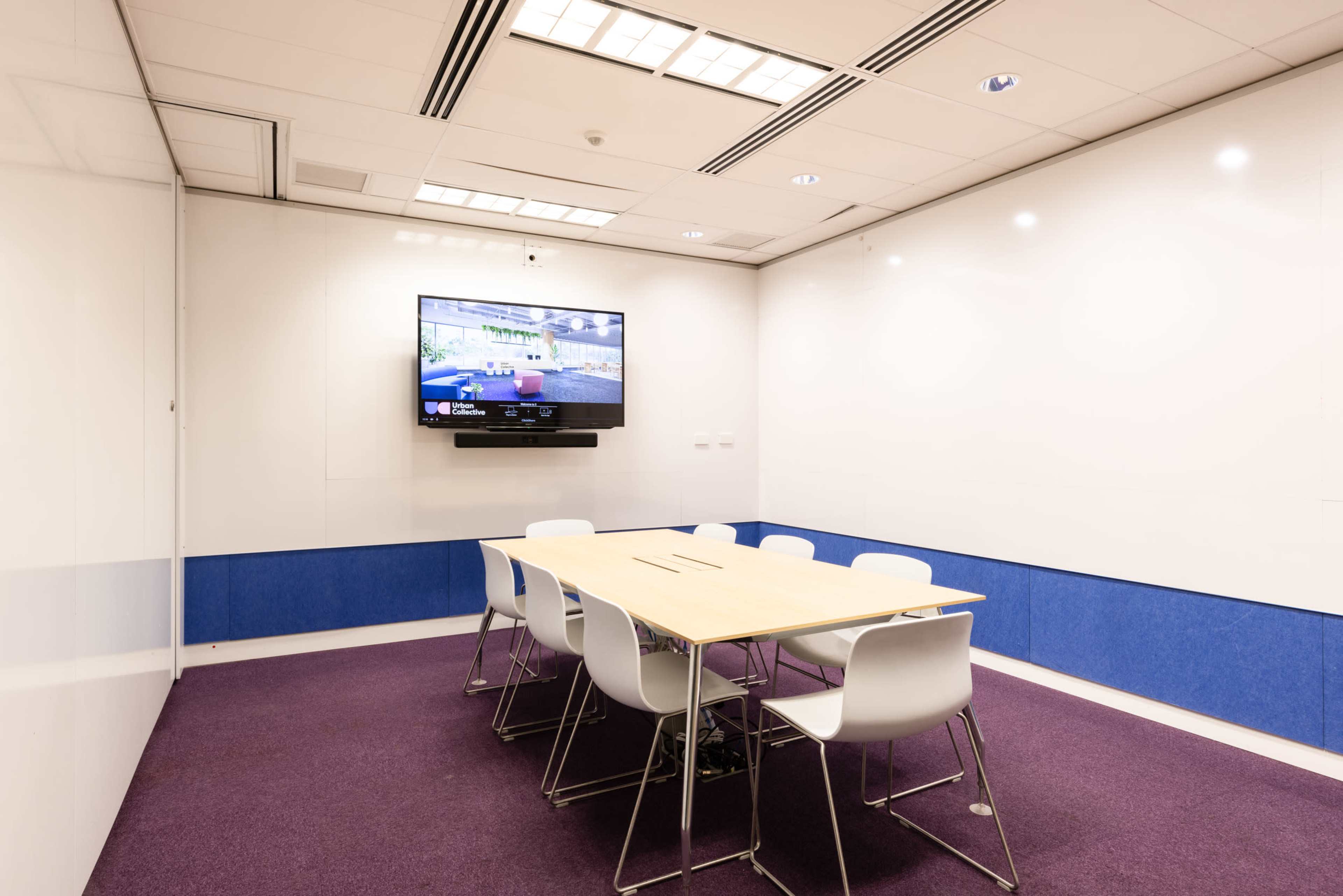 A modern conference room with a wooden table surrounded by white chairs and a wall-mounted television.