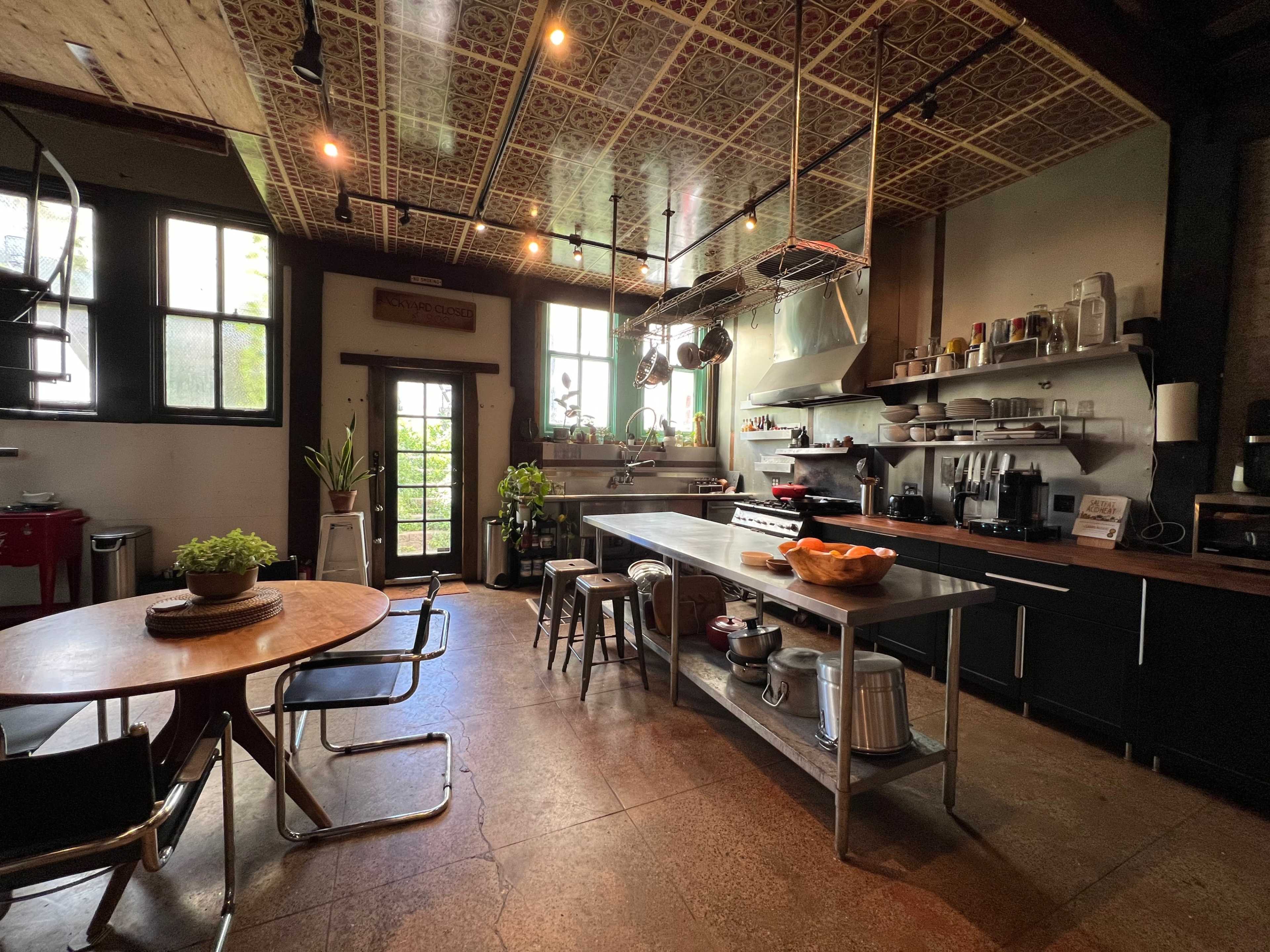 A modern kitchen with a large central island, wooden shelves filled with kitchenware, and a dining table with chairs, illuminated by natural light through multiple windows.