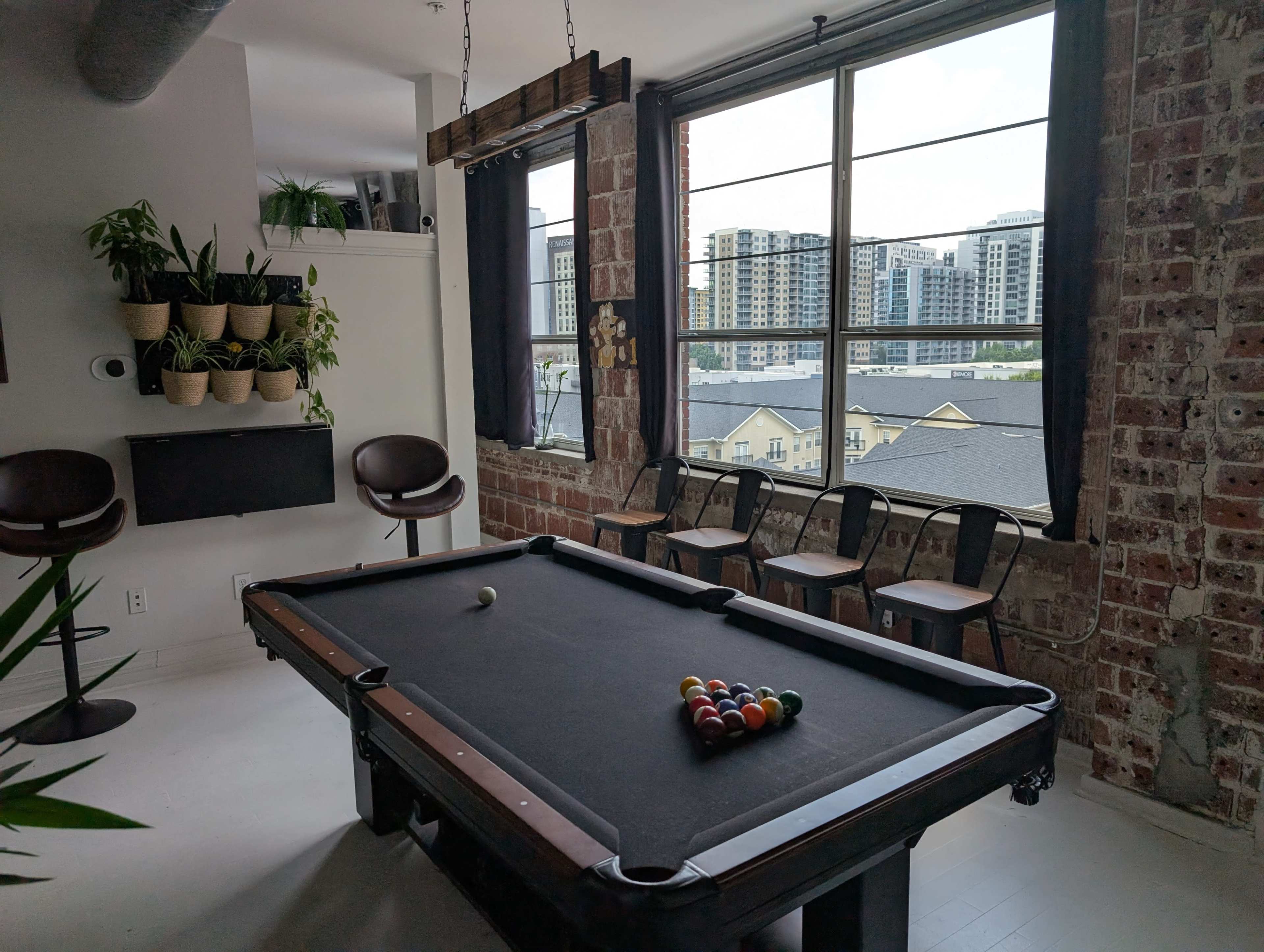 The image shows a room with a pool table in the center, exposed brick walls, and a large window overlooking modern buildings.