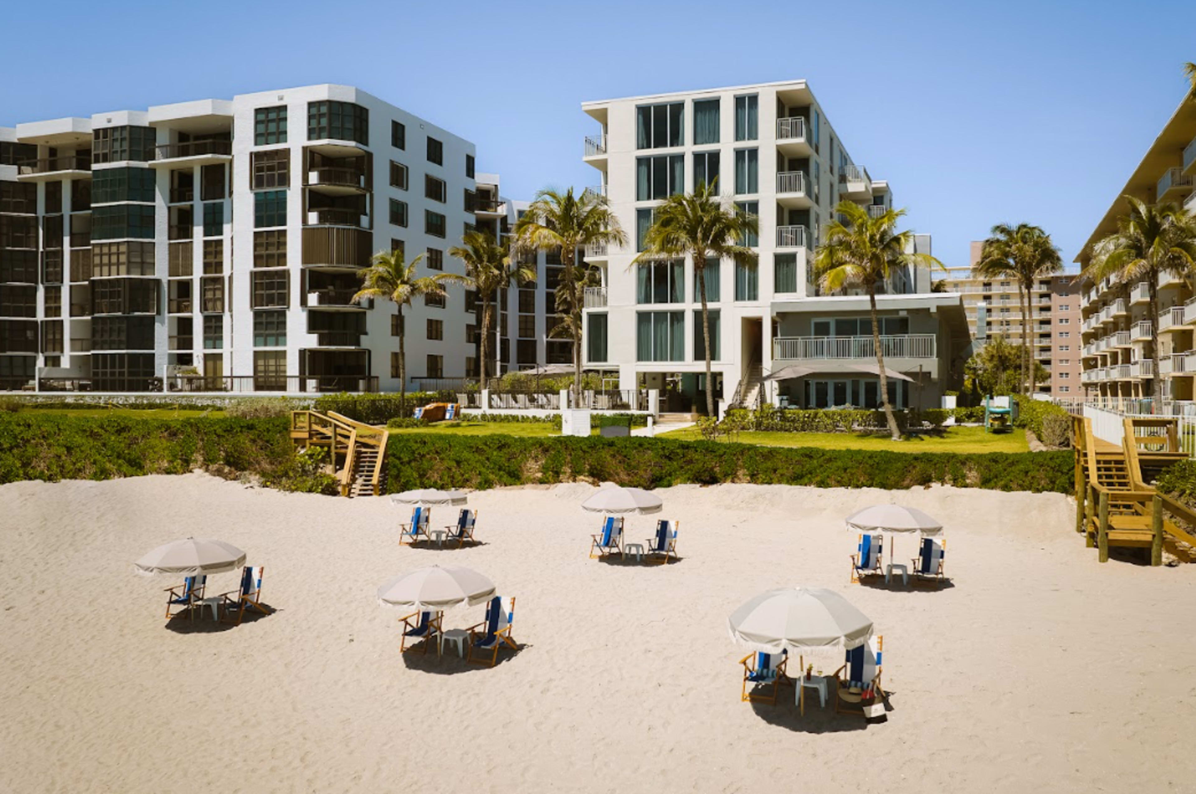 The image shows a beachfront scene with several beach umbrellas and chairs set up on the sand in front of modern buildings.
