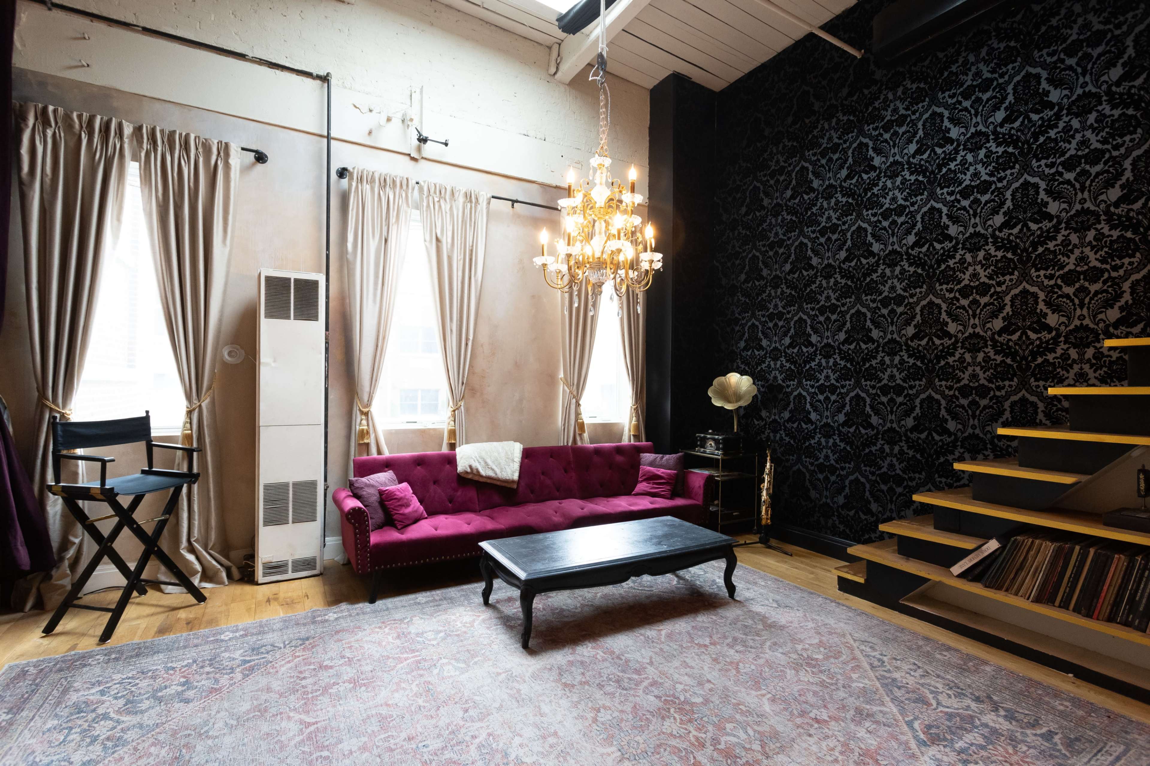 A spacious room features a velvet pink sofa, a chandelier, and a staircase leading to a darker wall adorned with a decorative pattern.