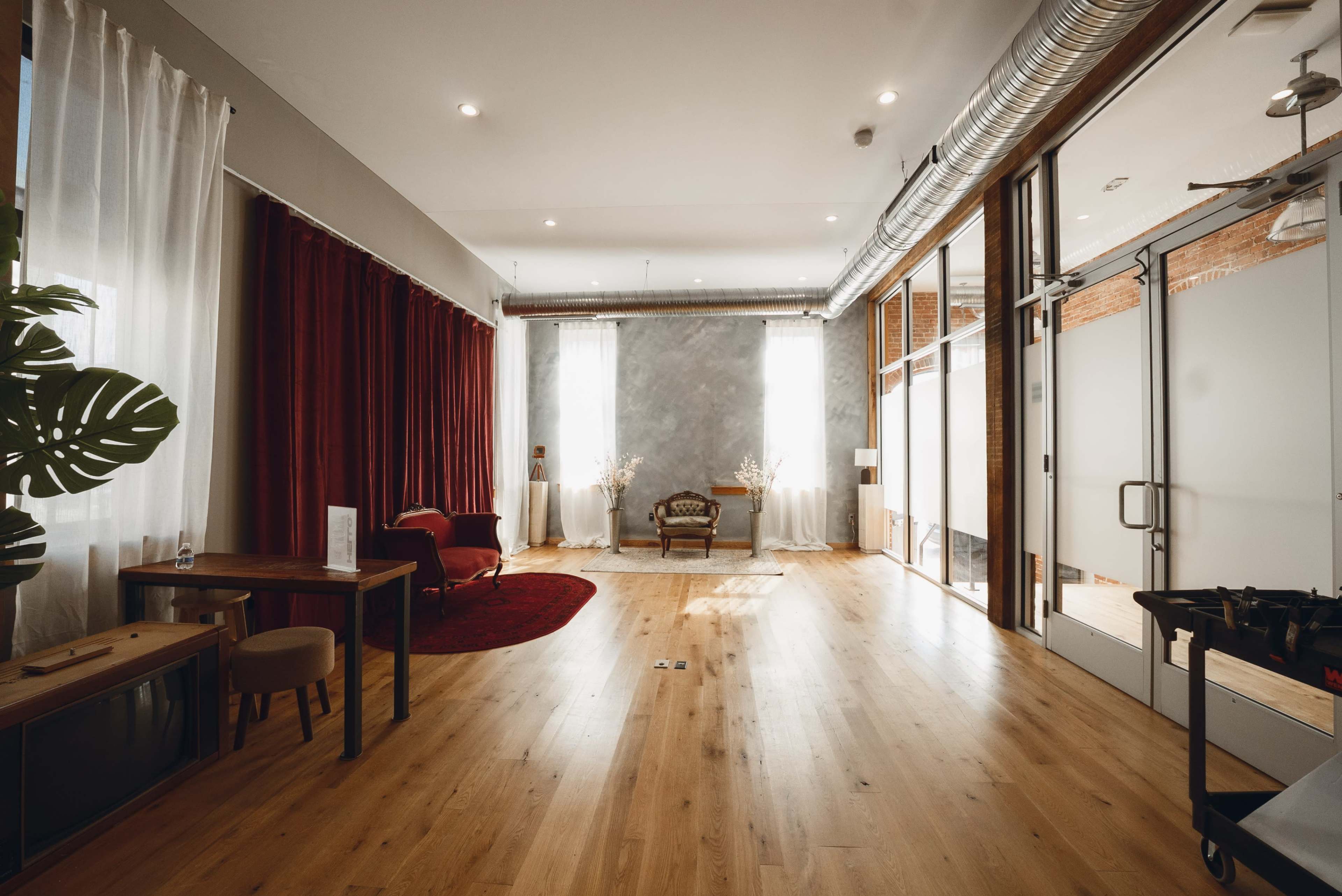 The image shows a minimalist room with wooden flooring, large windows with sheer curtains, and a combination of red drapes and a gray wall.