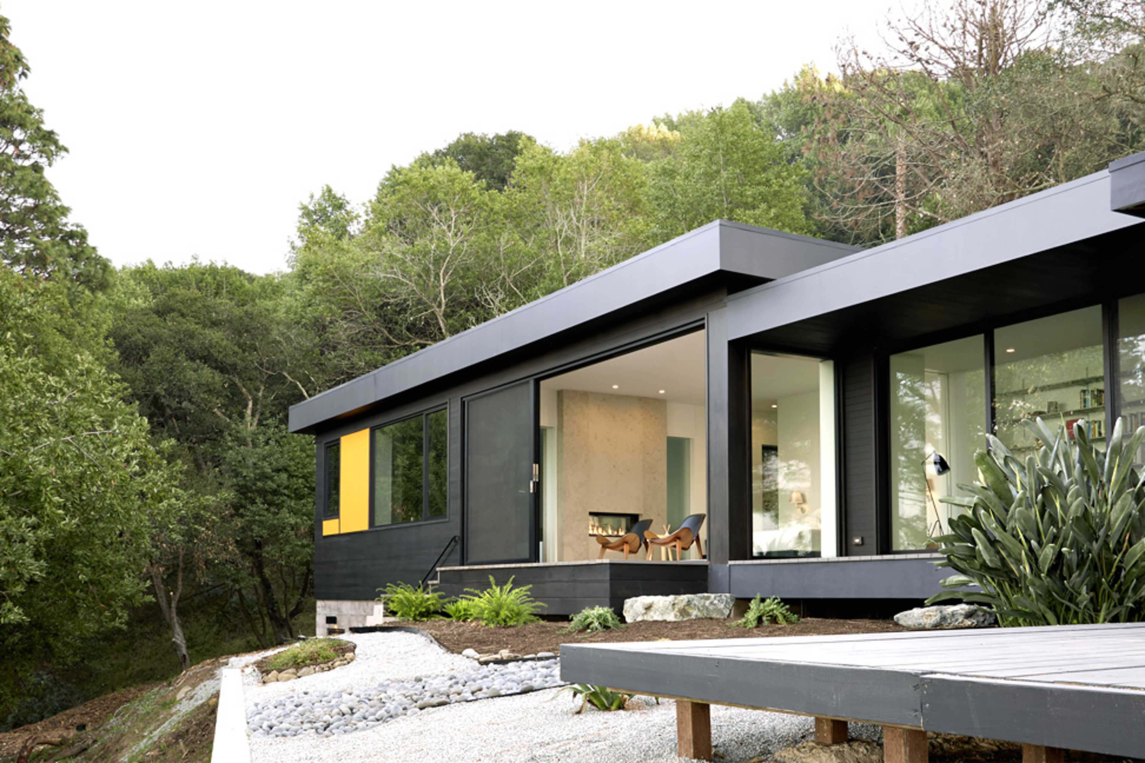 A modern house with large windows and a contrast of black siding and yellow accents overlooks a landscaped yard and surrounding trees.