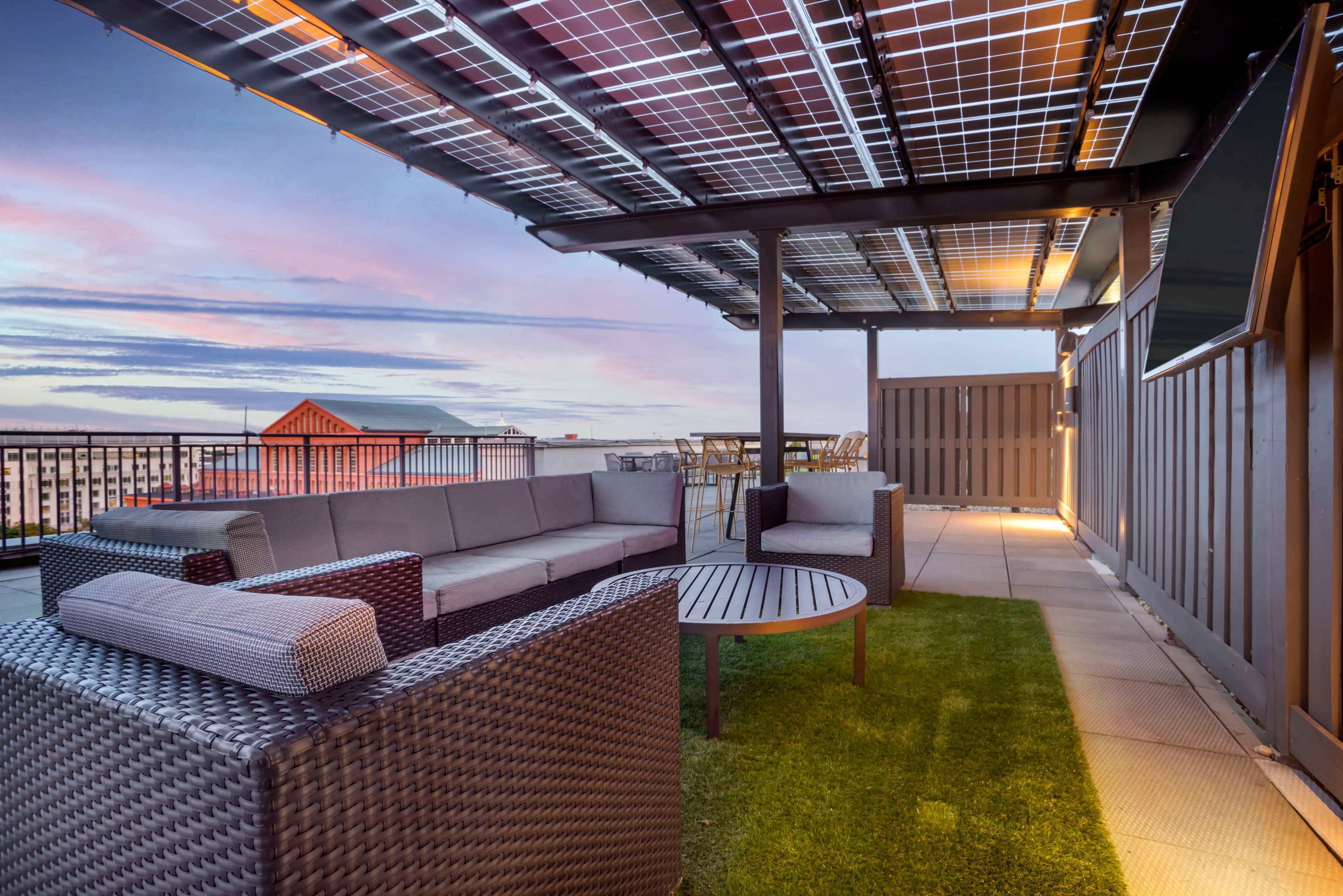 A rooftop patio features a sectional sofa, a round table, and a grassy area under a solar panel-covered pergola, with a view of the sky at dusk.