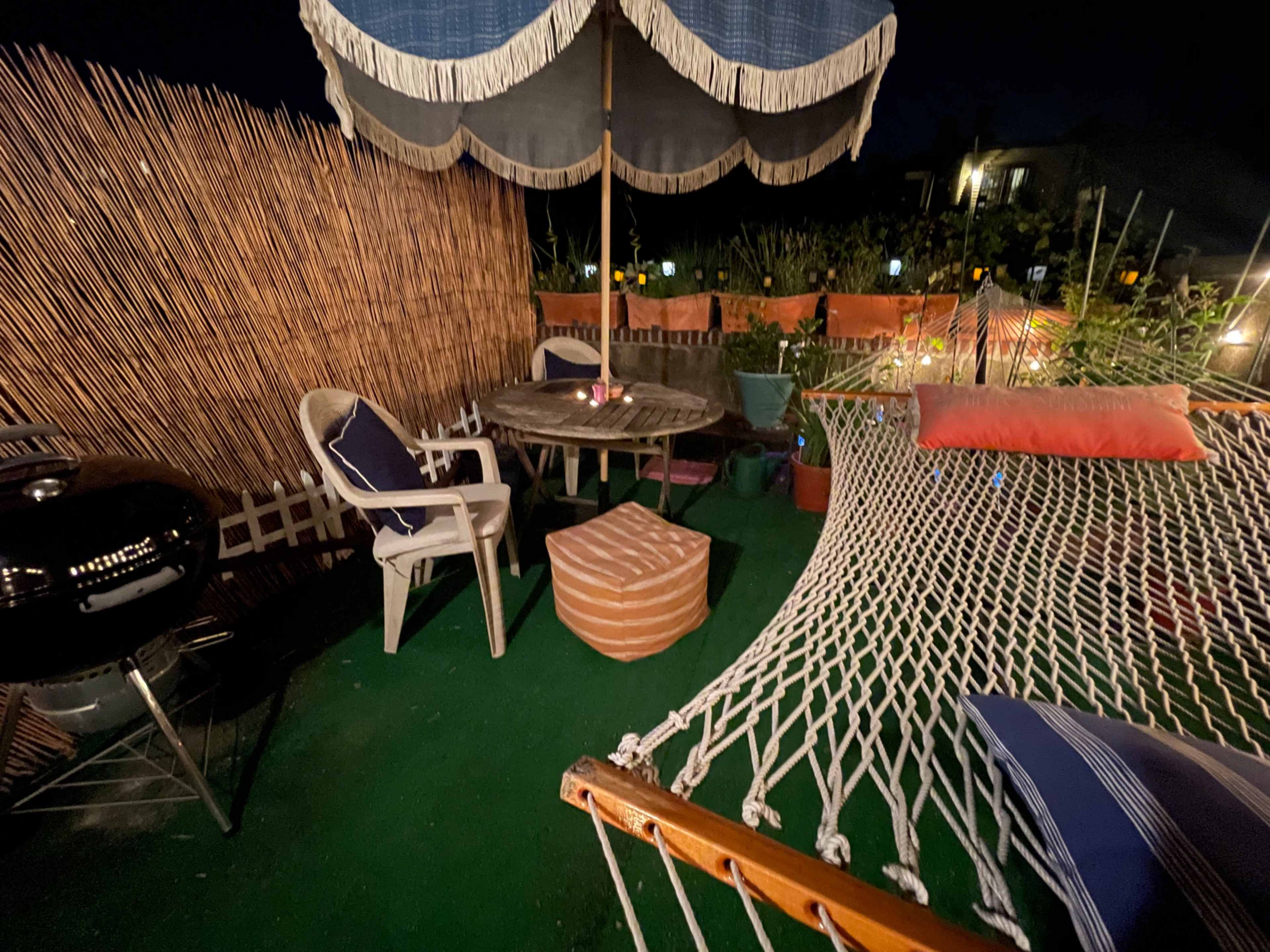 A cozy outdoor patio features a hammock, a circular table with chairs, a grill, and ambient lighting surrounded by bamboo fencing.