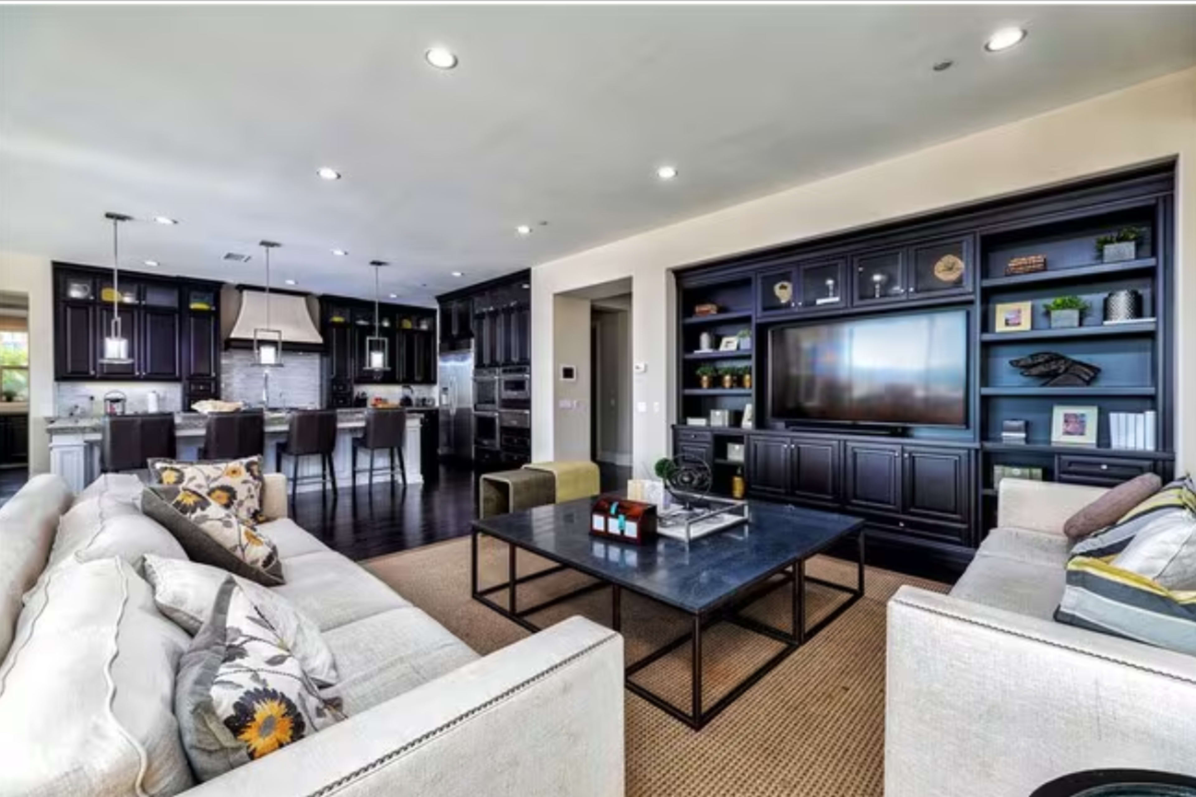 The image shows a modern living room with two sofas facing a black coffee table, a built-in entertainment center, and a view of a kitchen area in the background.