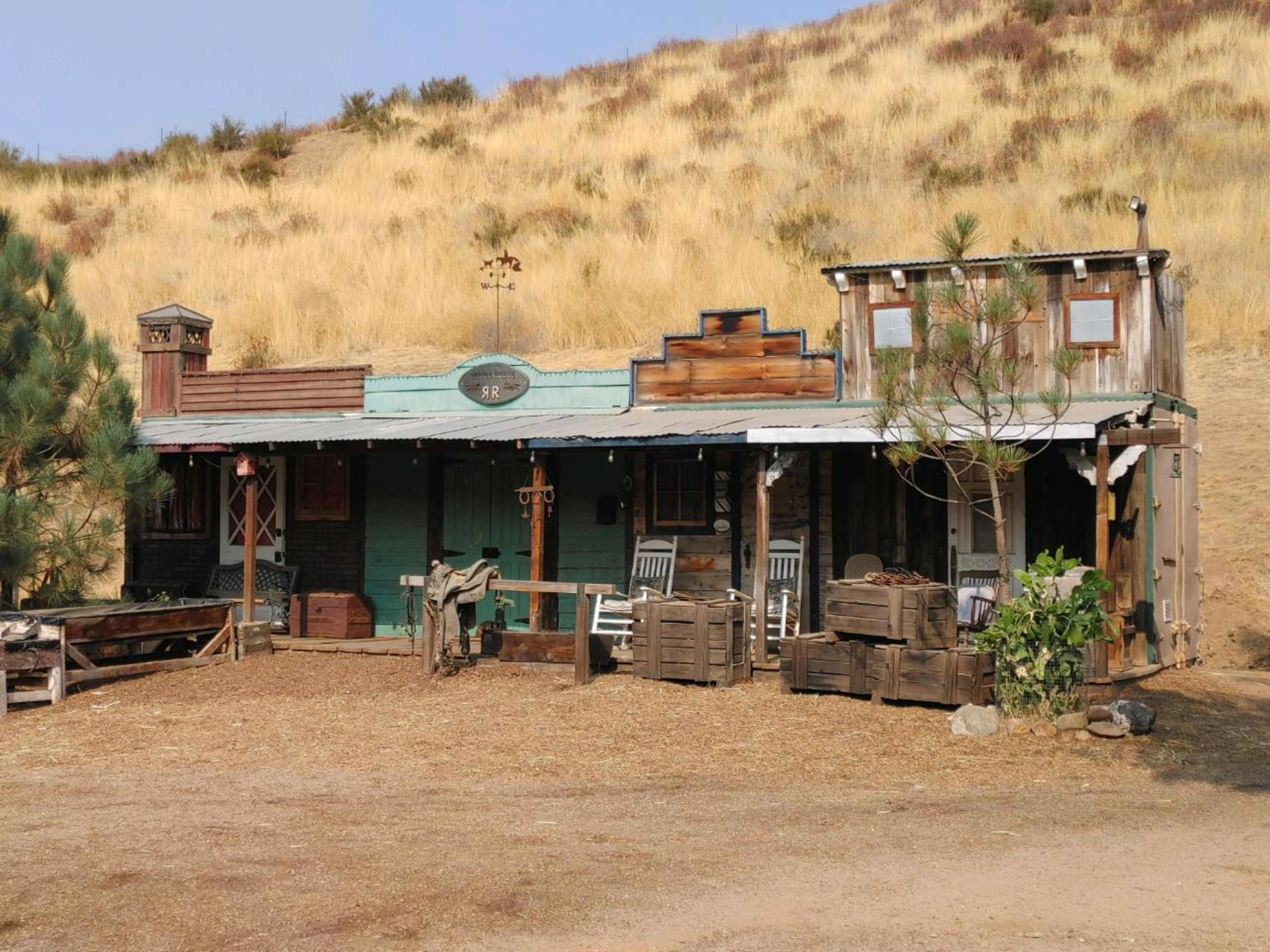 The scene features a rustic, western-style building with a wooden facade, a porch with rocking chairs, and crates arranged outside, set against a hillside covered in dry grass.