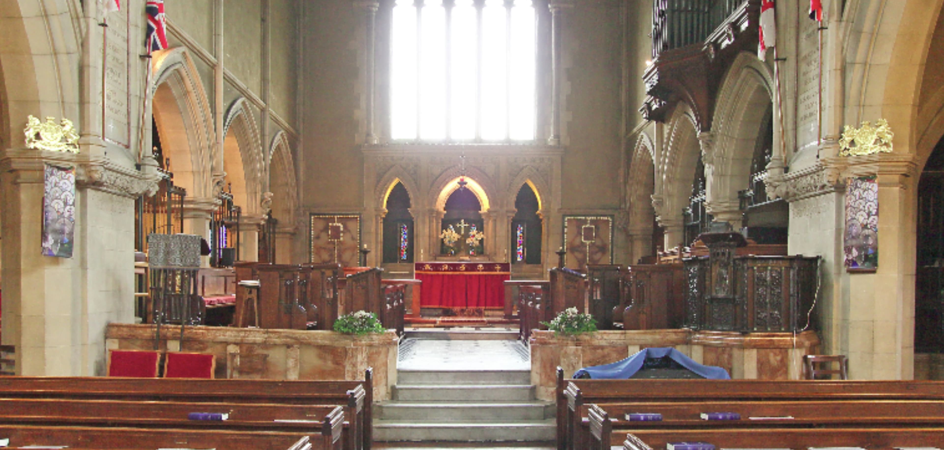 The interior of a church features wooden pews, stone arches, and a prominent altar with red decorations at the front under tall windows.