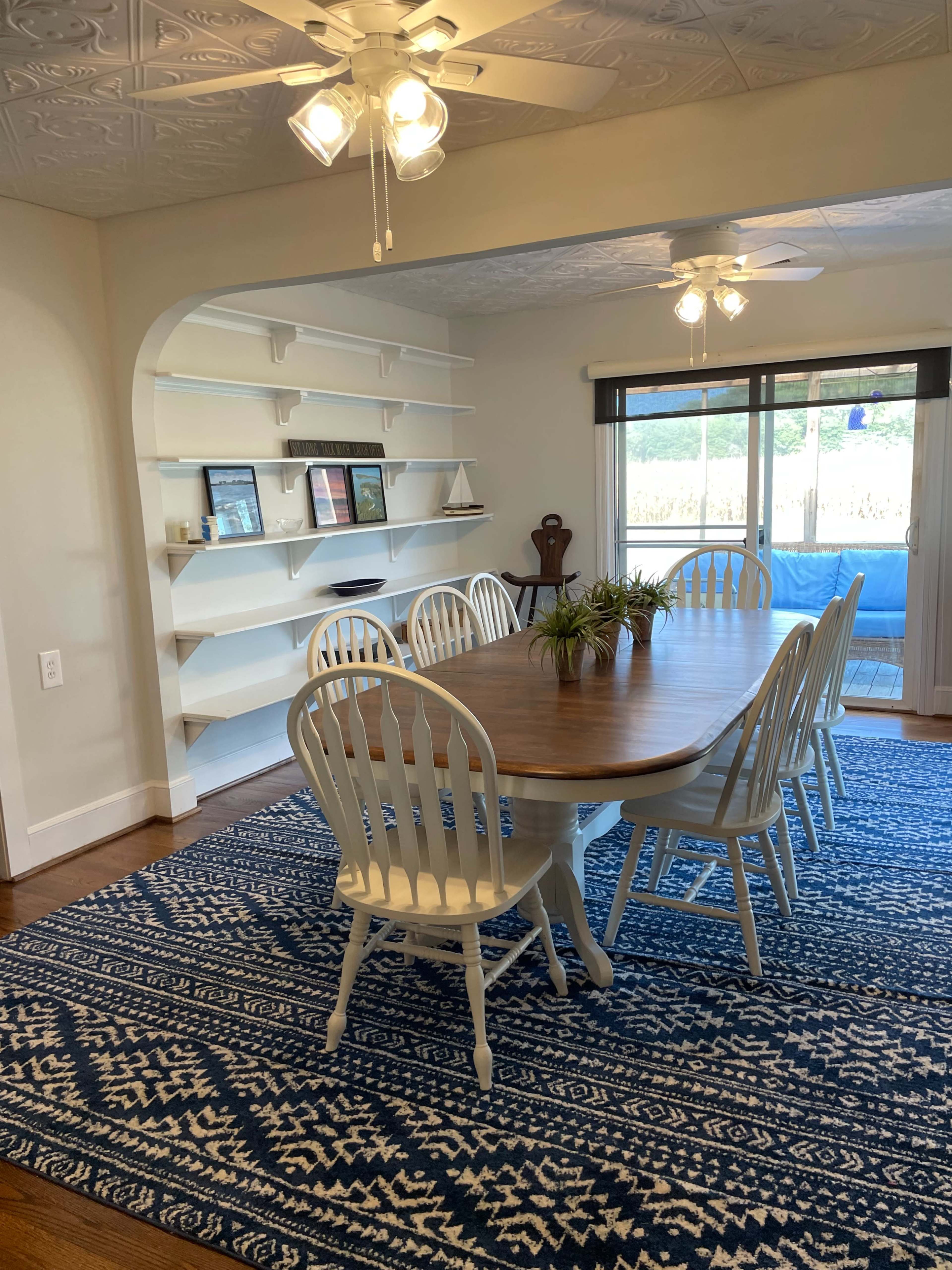 A dining room with a large wooden table surrounded by white chairs on a blue patterned rug, and a set of shelves on the wall displaying framed pictures and decor.