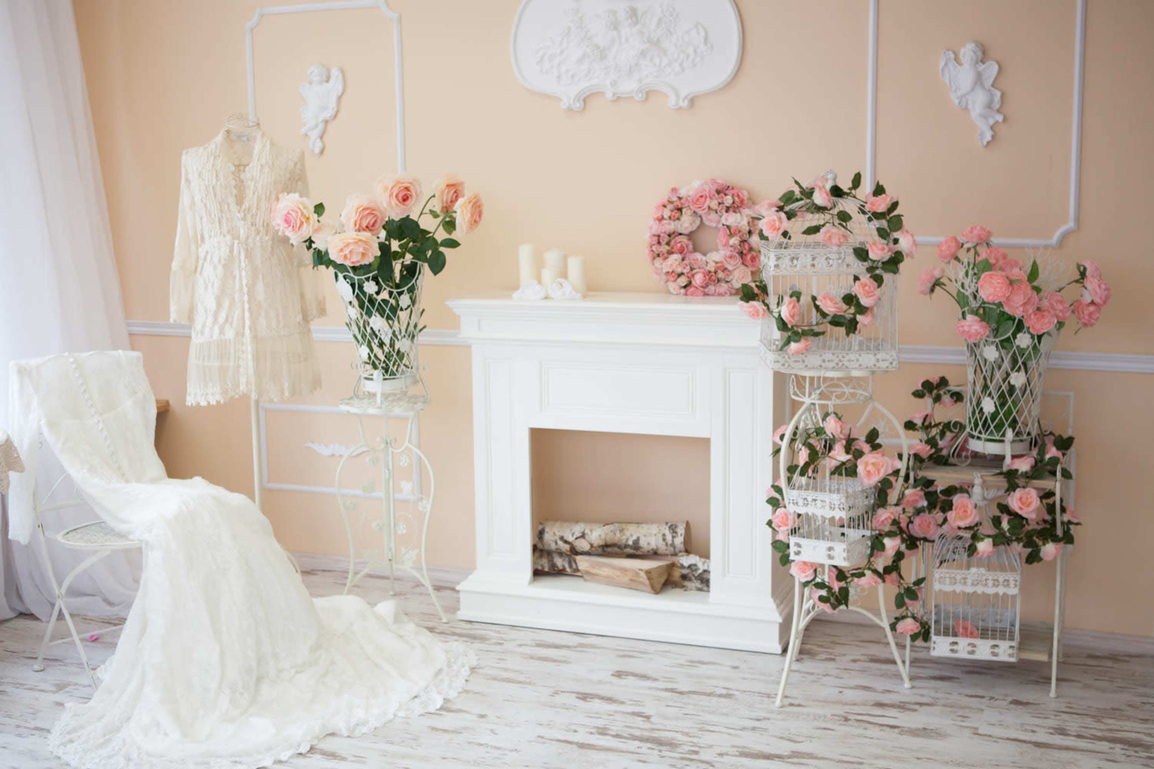 The image features a softly decorated room with a white fireplace, adorned with flowers in decorative stands, a lace dress on display, and a white chair.
