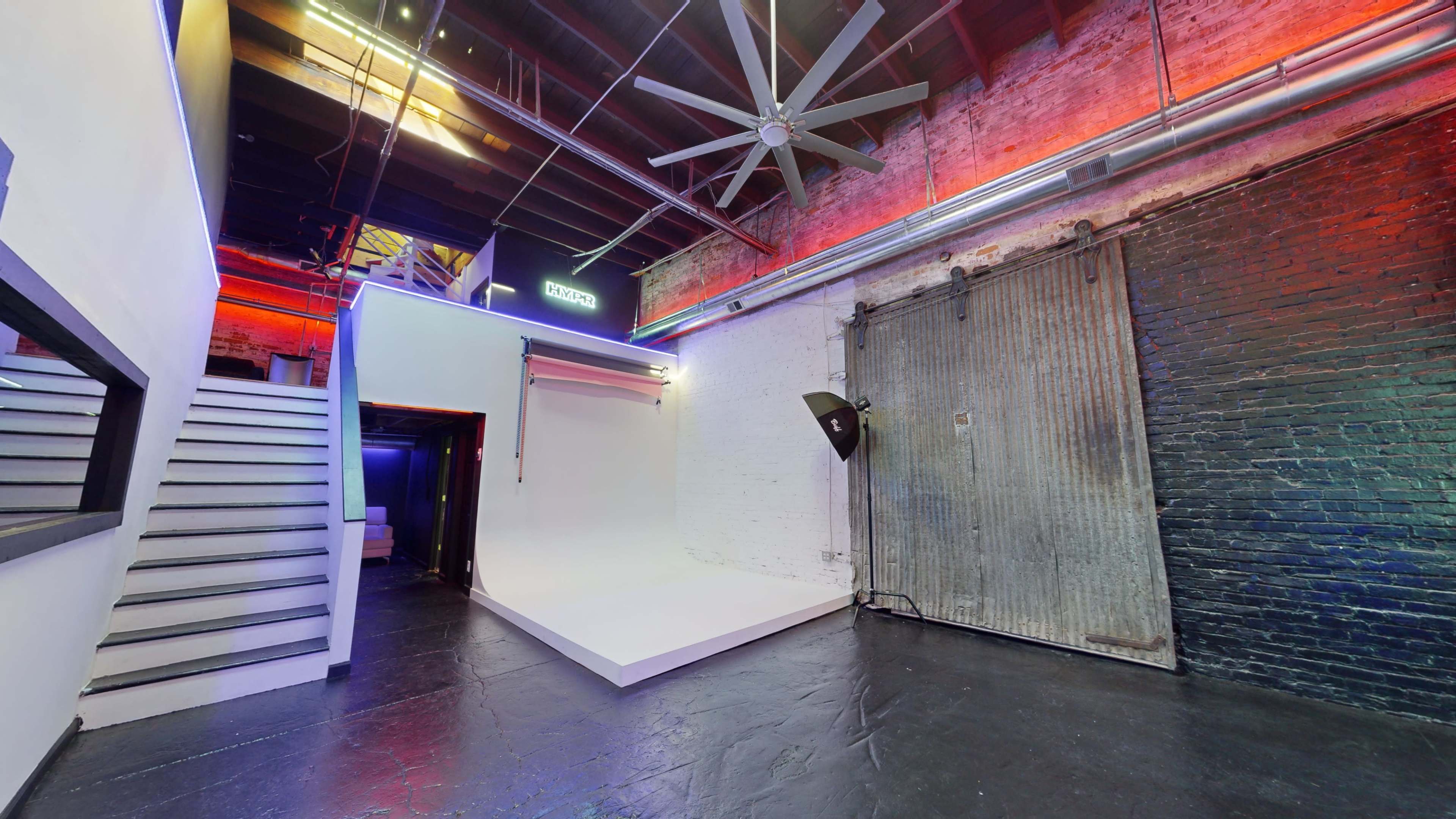 An industrial-style studio space with a white backdrop, large ceiling fan, exposed brick walls, and a set of stairs leading to an upper level.