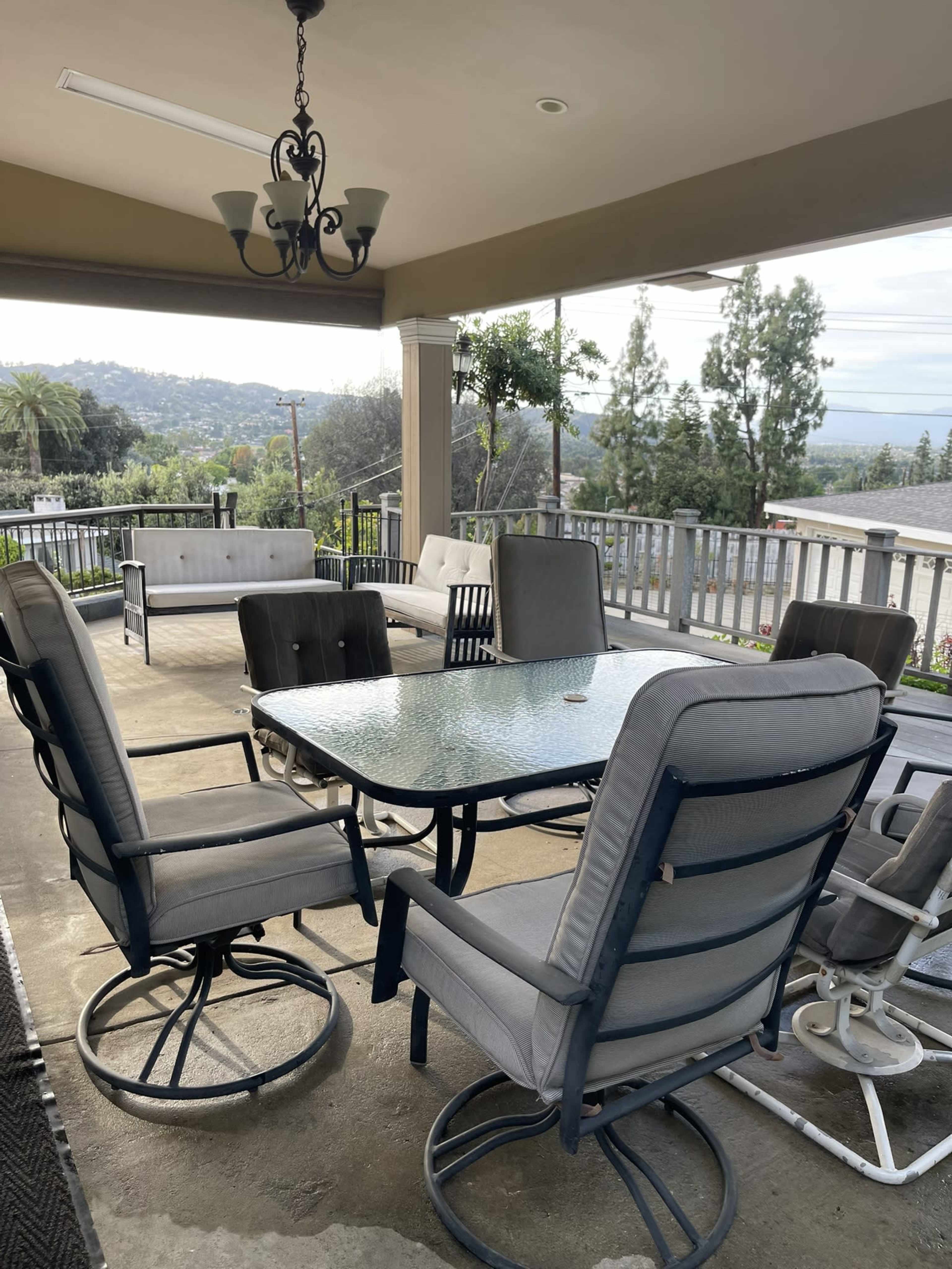 The image depicts a covered patio with a table and chairs, overlooking a scenic view of trees and hills in the background.