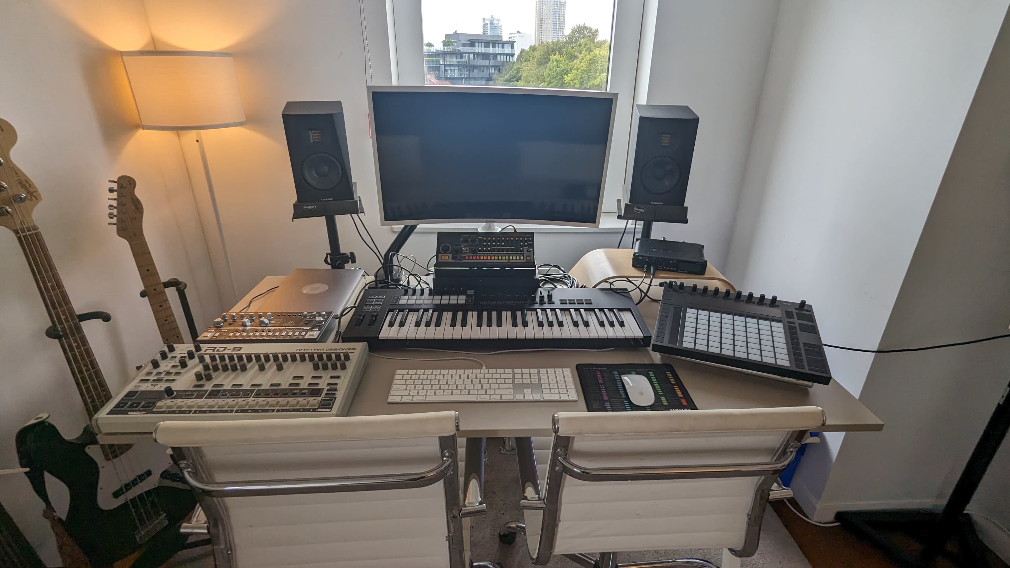 The image shows a music production setup featuring a keyboard, audio interface, drum machine, computer monitor, and studio monitors on a desk with two chairs.