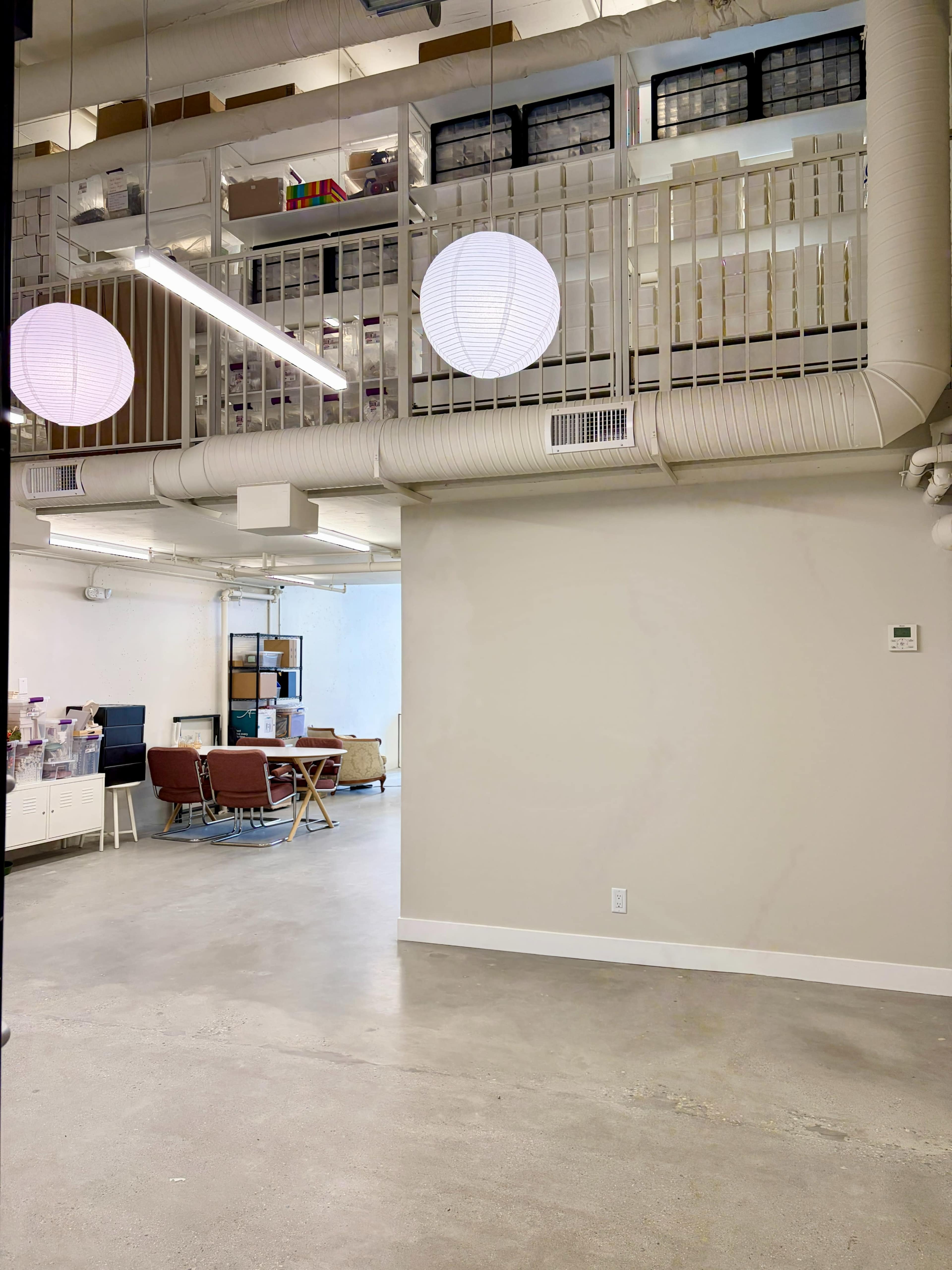 The image shows a spacious office area with a light-colored wall and polished concrete floor, featuring a seating area with red chairs and a balcony above lined with storage bins.
