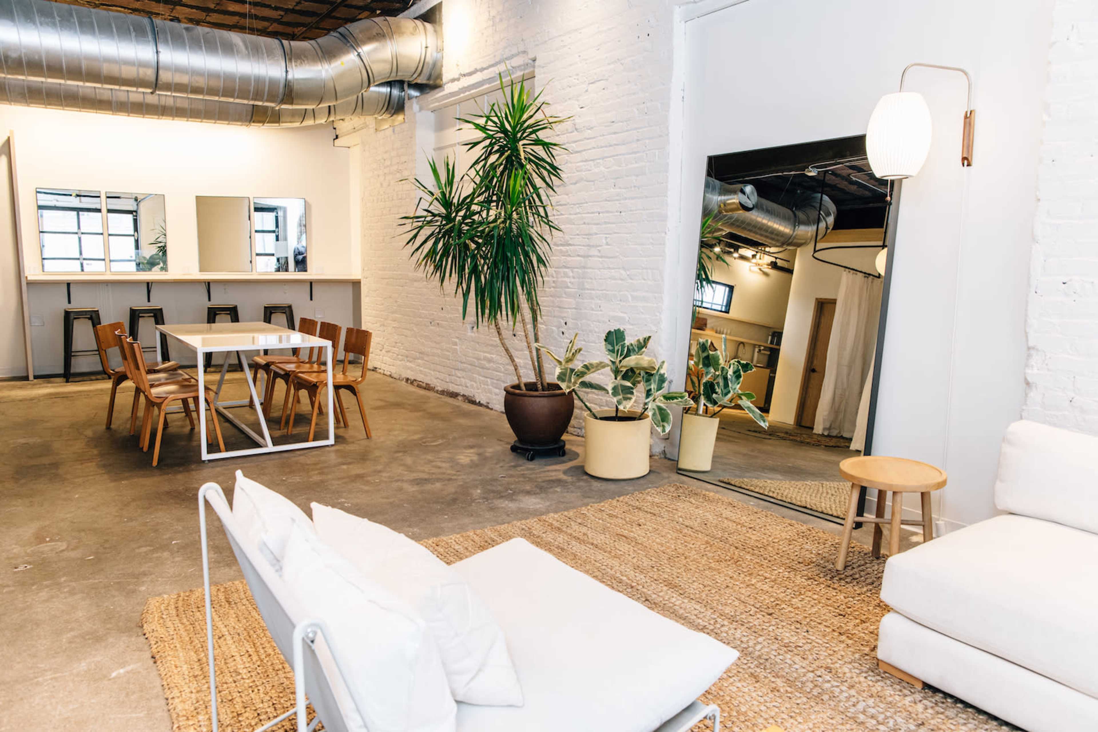 The image shows a spacious, industrial-style room featuring a seating area with a white sofa, a dining table, wooden chairs, and various potted plants.