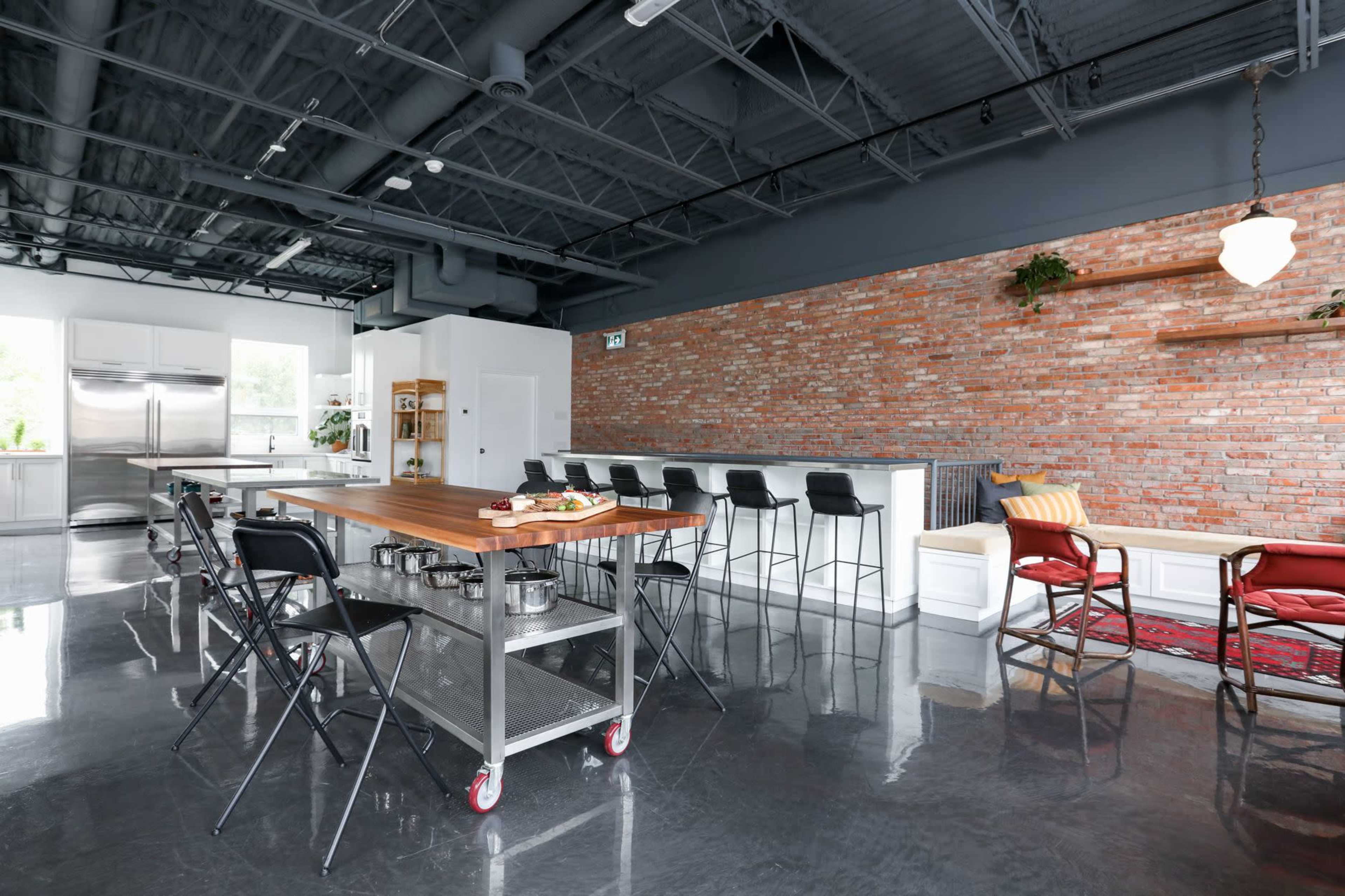 A modern kitchen with a large wooden island, metal stools, and a brick wall, featuring stainless steel appliances and a minimalistic design.