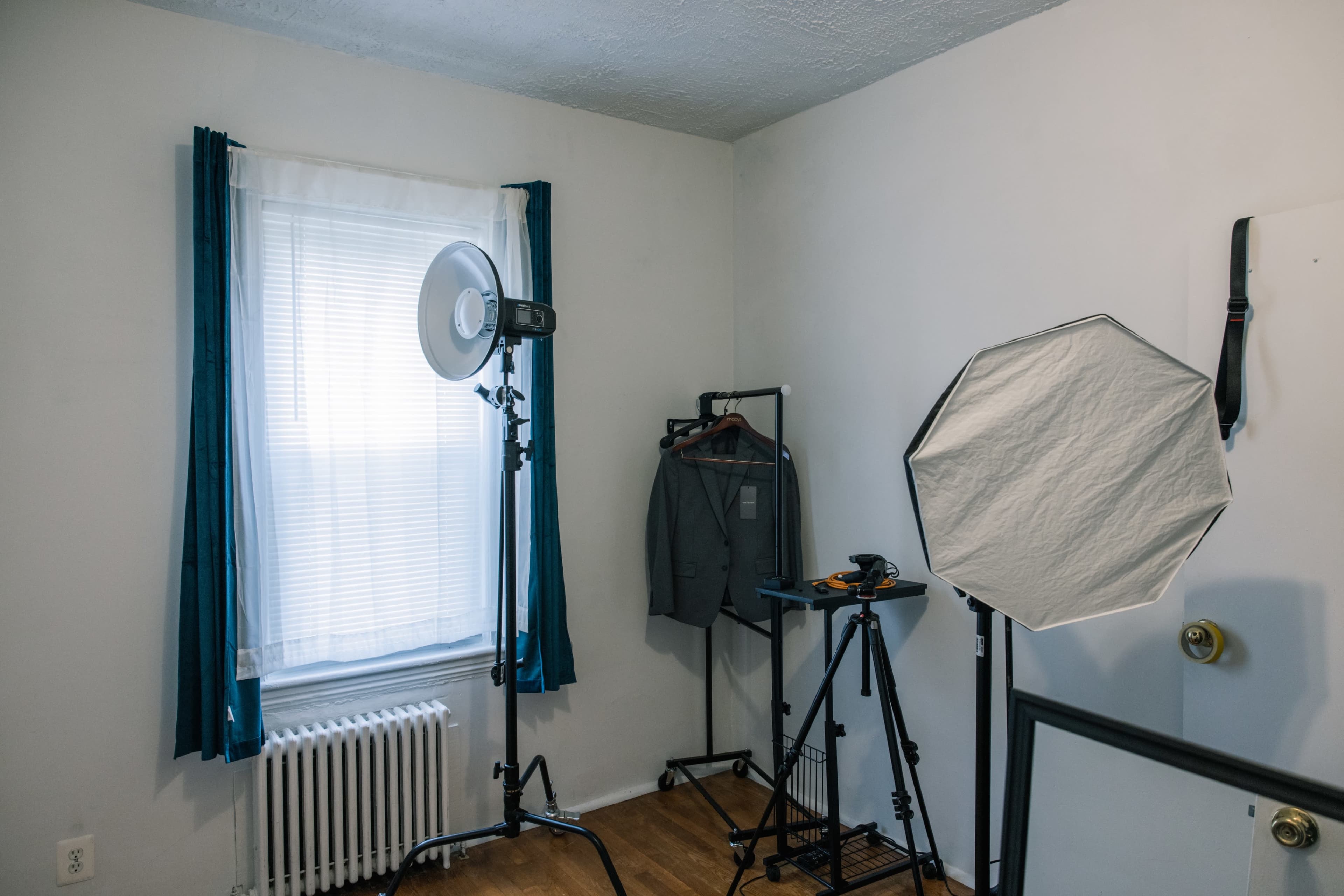 A room with a window, a suit hanging on a clothing rack, a light stand with a softbox, and a tripod.