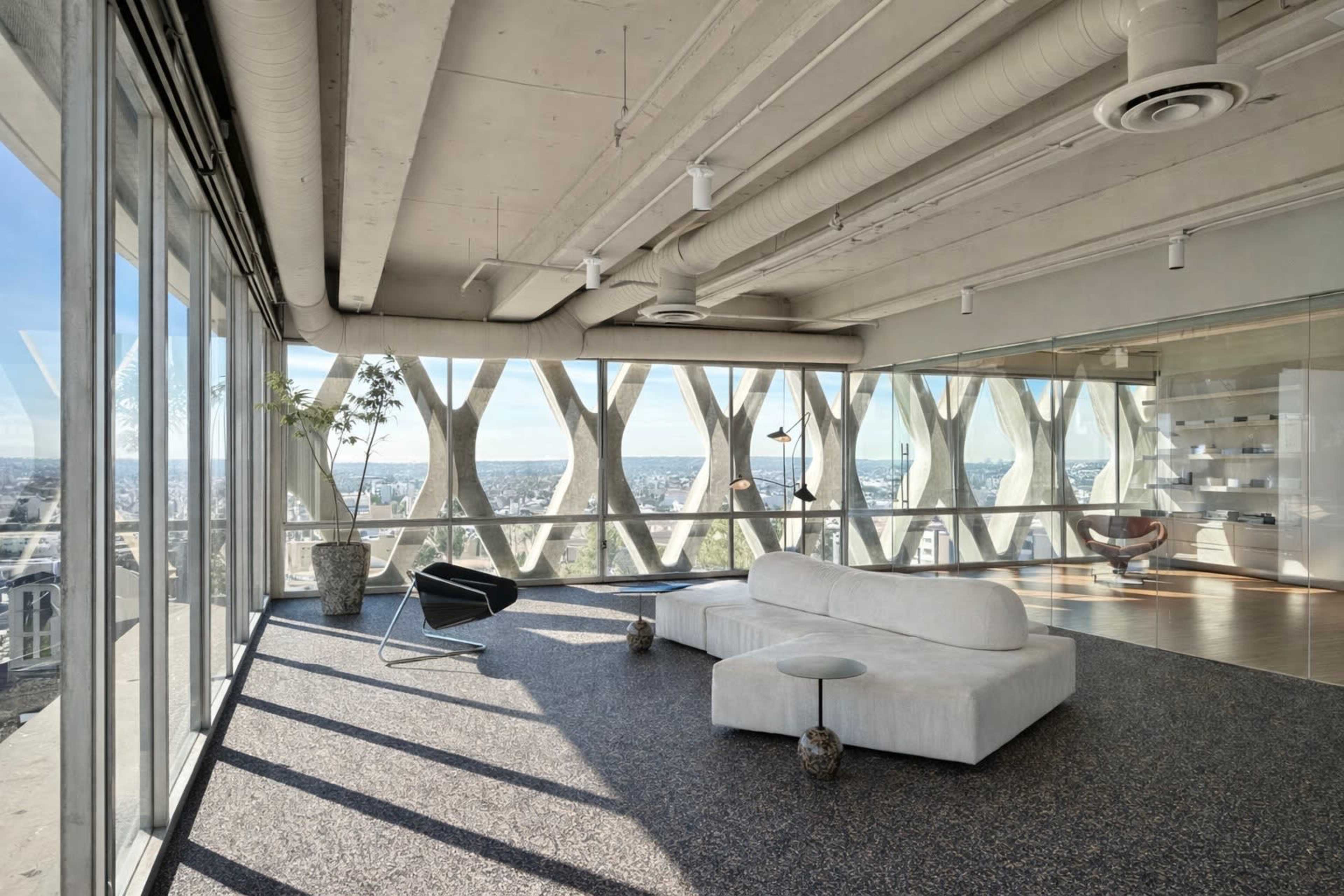 The image shows a modern, spacious interior with large windows offering a panoramic view of the city, featuring minimalist furniture and exposed concrete ceilings.