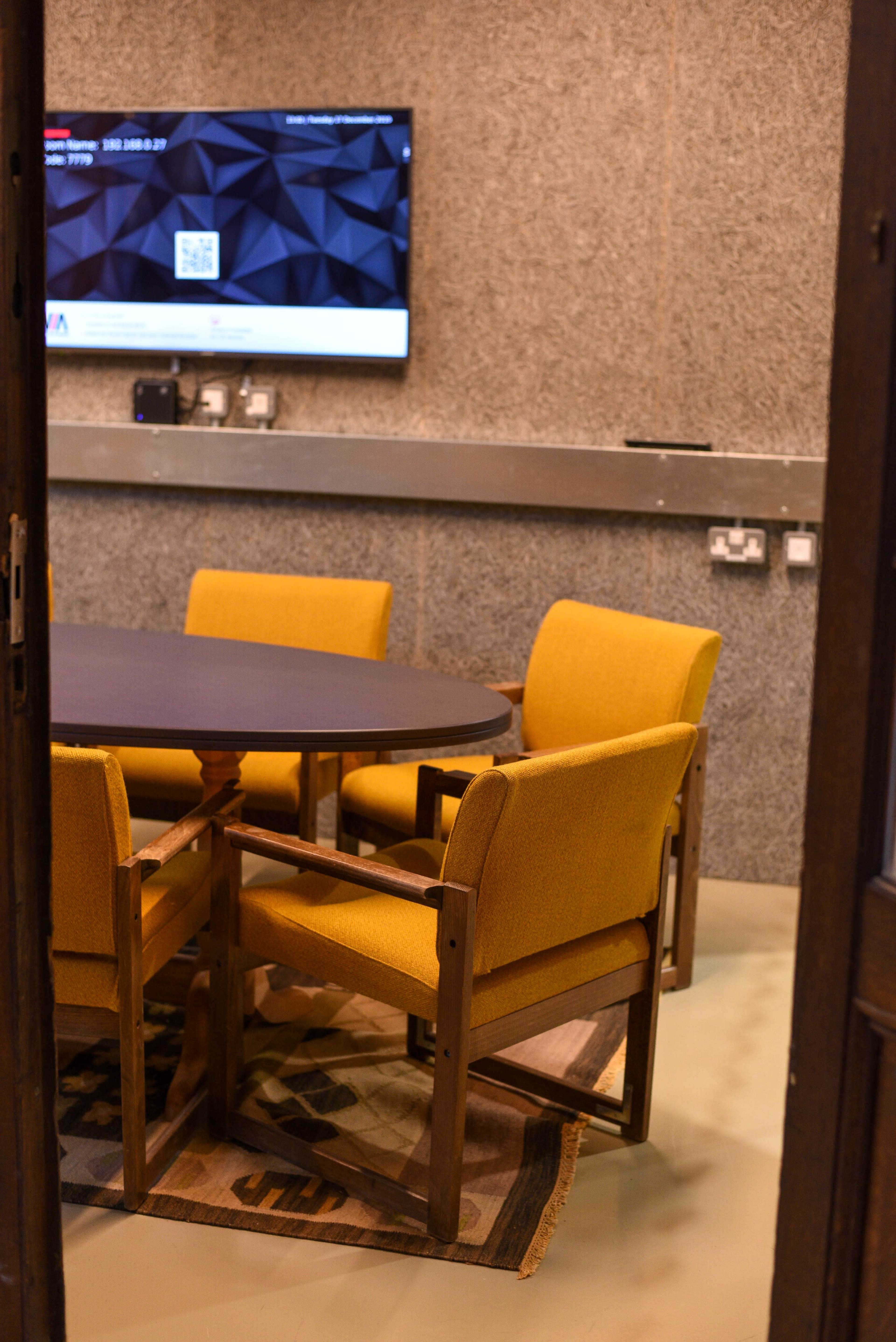 The image shows a meeting room with a round table surrounded by four yellow chairs and a television mounted on a gray wall.