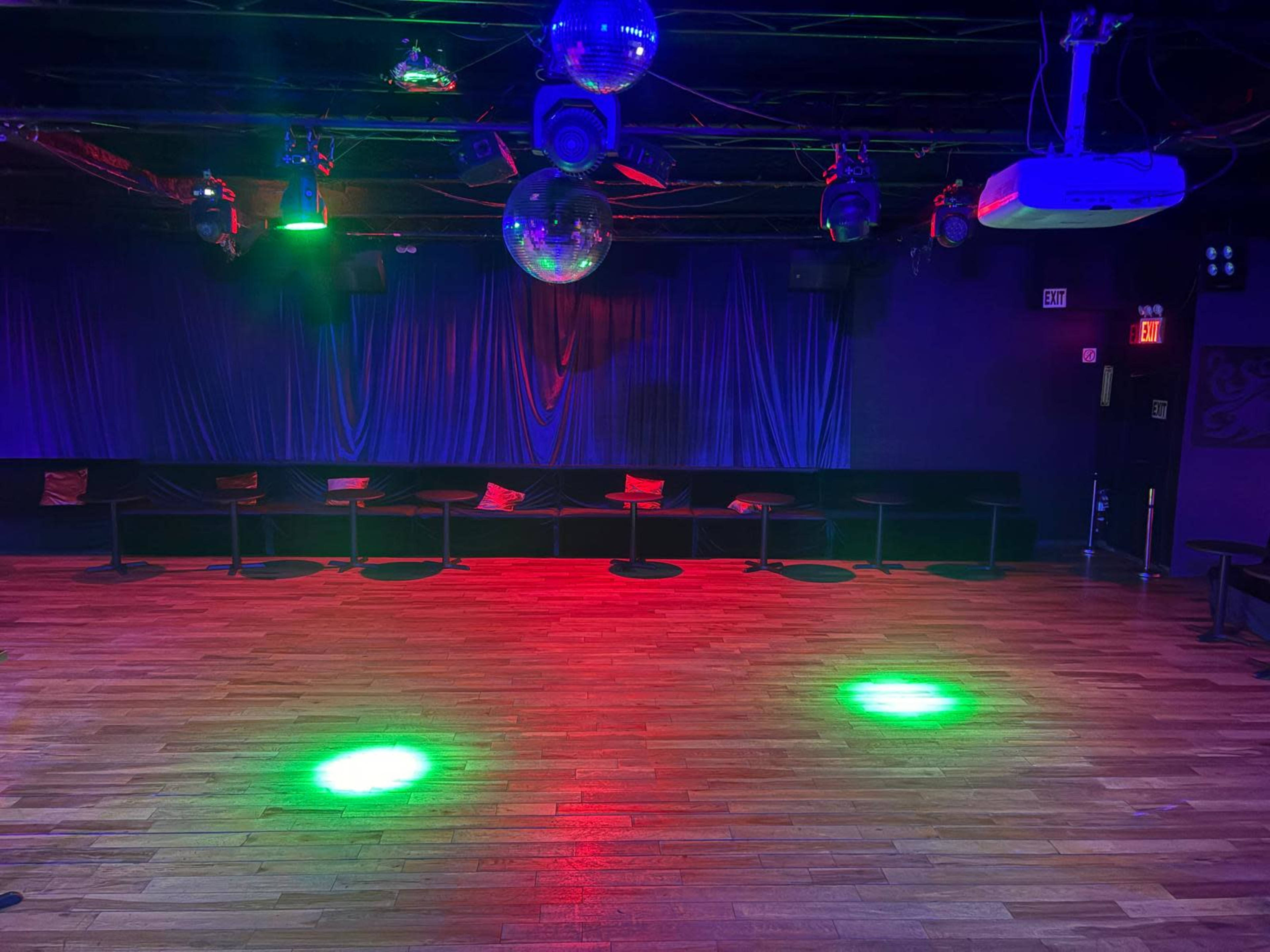 The image shows an empty dance floor with wooden flooring, illuminated by green and red lights, and adorned with disco balls hanging from the ceiling.